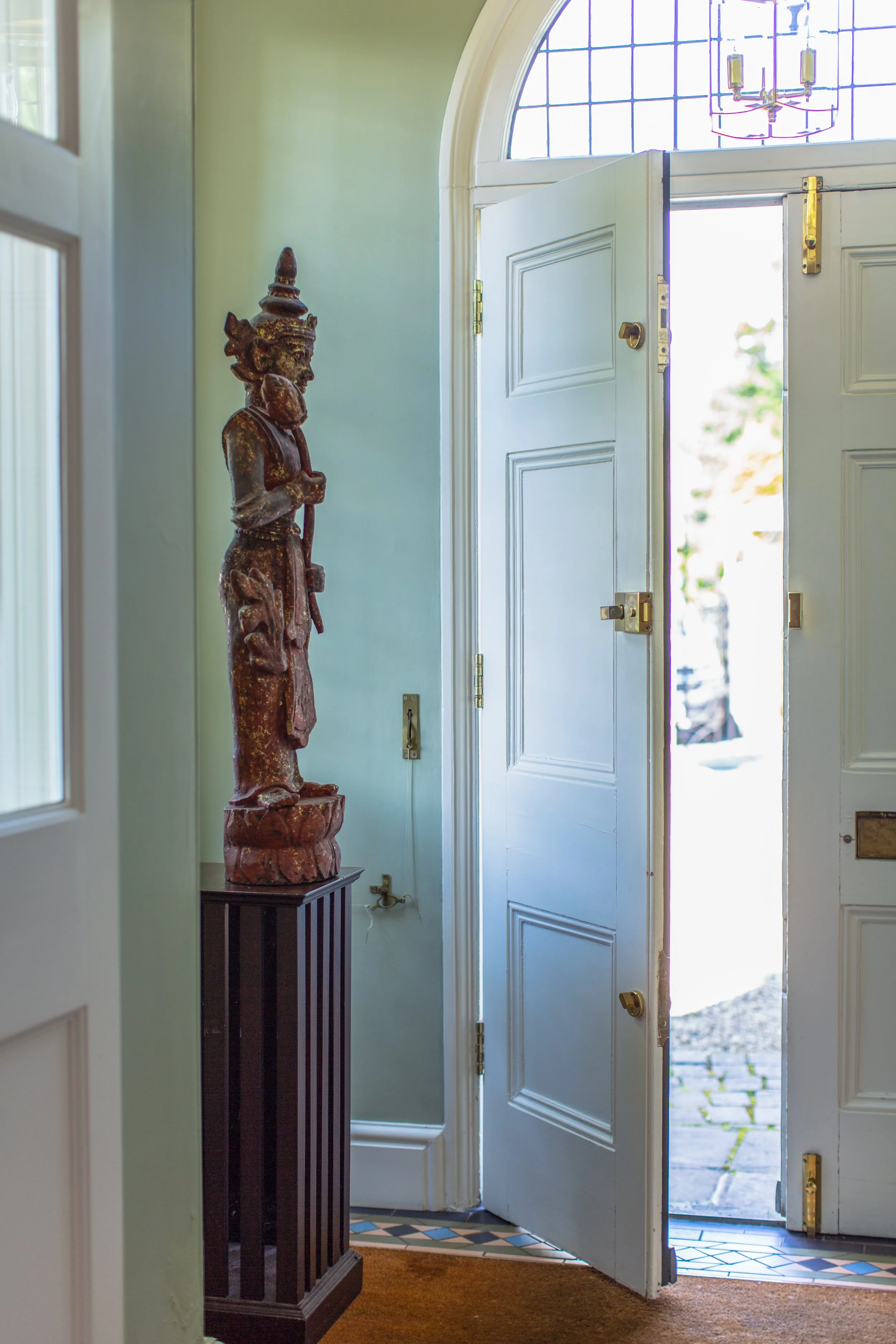 Open front door of a house with a decorative wooden statue of a figure wearing a headdress inside, standing on a dark wooden pedestal near the door, with light streaming outside.