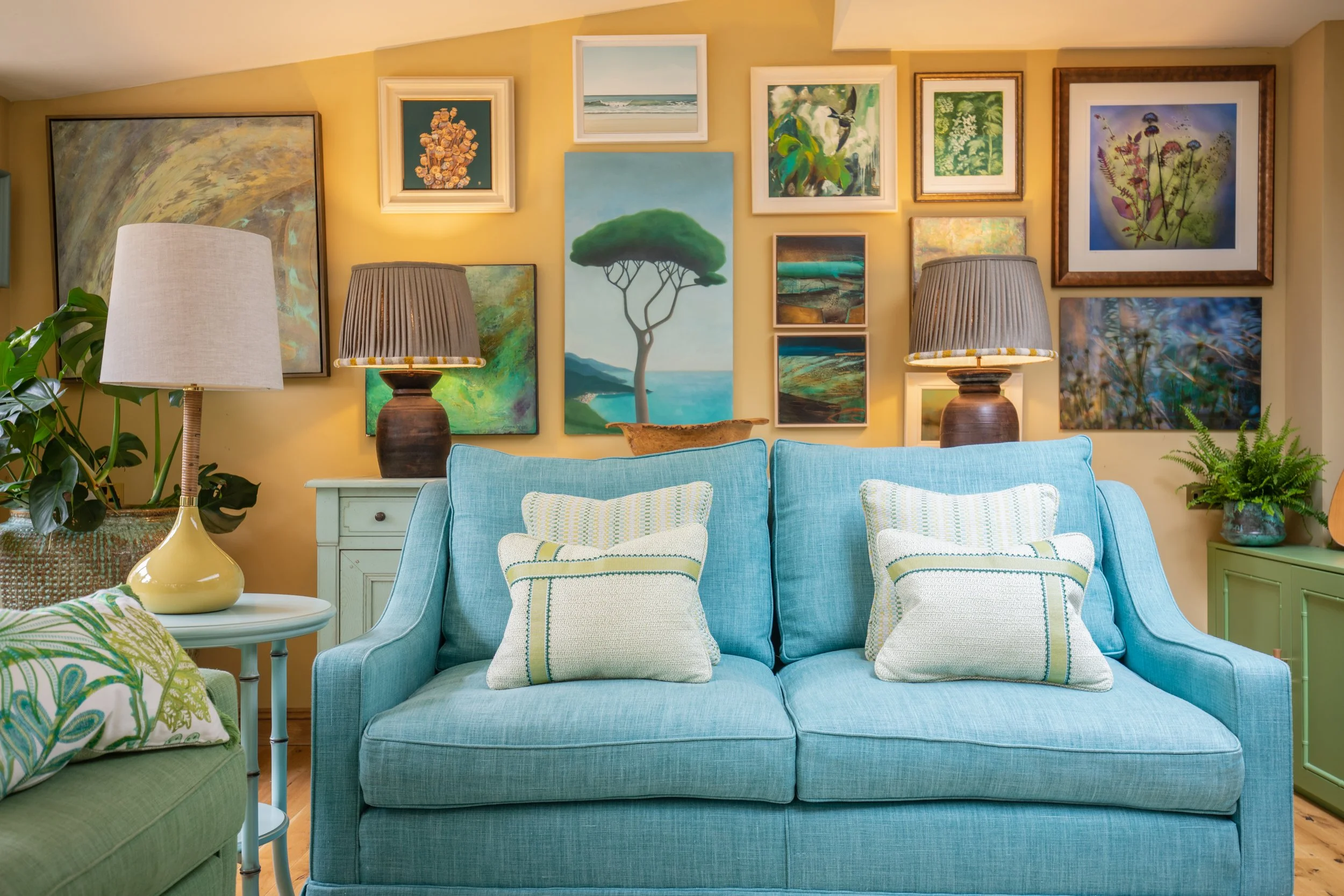 Living room with a blue couch, colorful pillows, two lamps, and a gallery wall of framed artwork on a yellow wall. There are plants on the sides.
