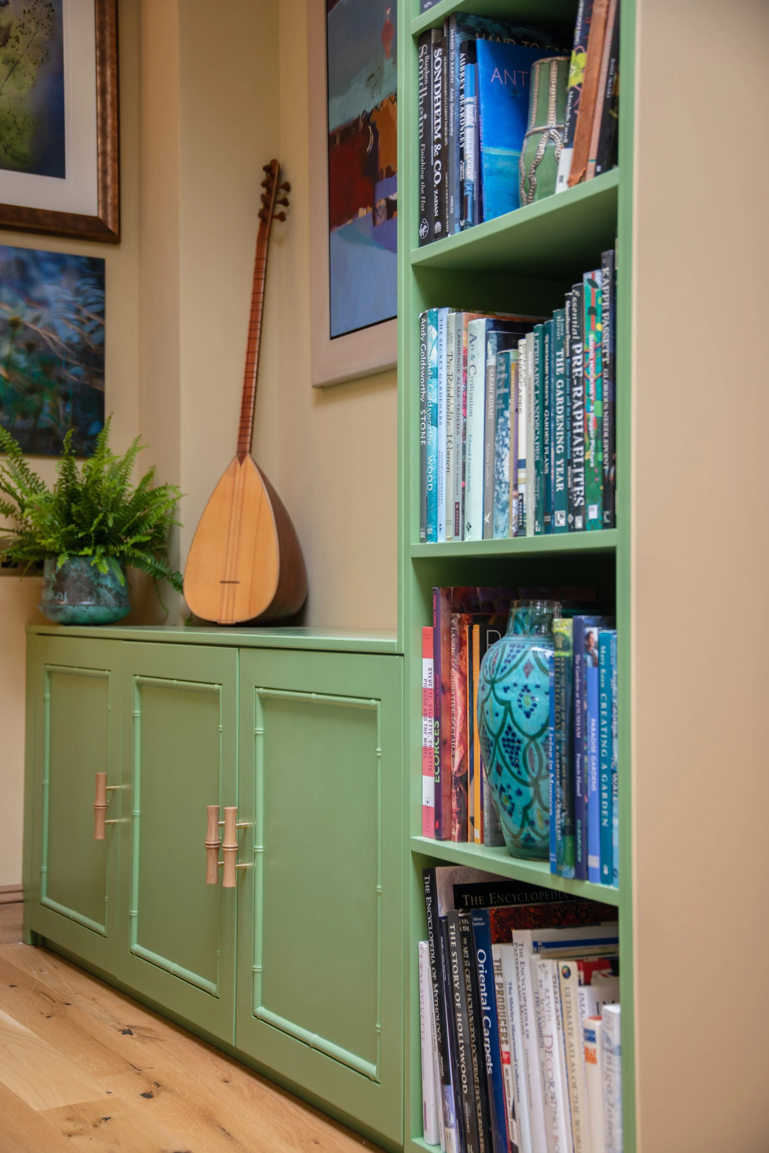 A green bookshelf filled with books, a decorative vase, a turquoise glass vase, and a potted fern on top of a green cabinet, with framed artwork on the beige wall behind.