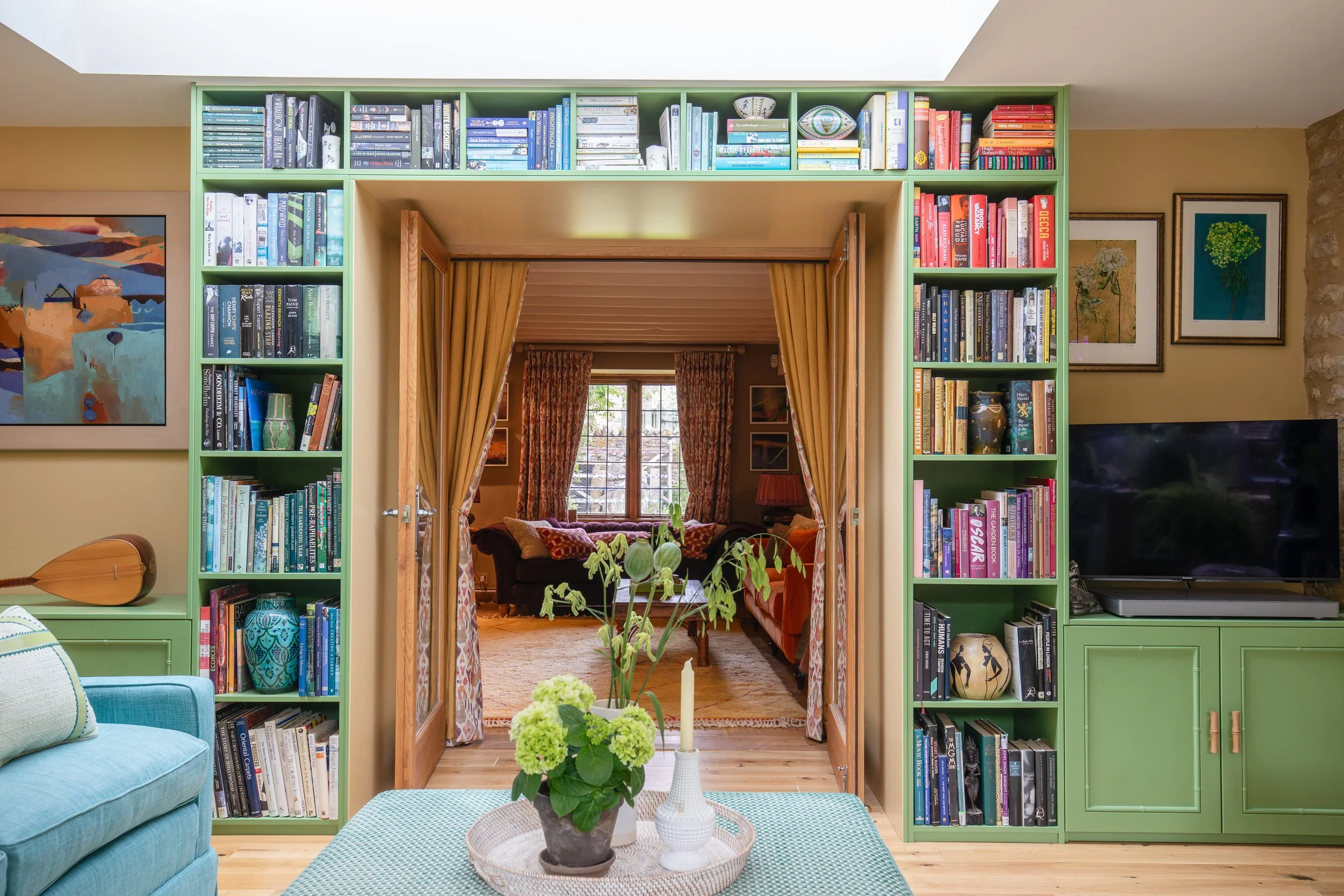 Living room with bright green bookcase filled with books, a teal sofa with pillows, a round coffee table with a potted plant and candles, and a doorway with curtains leading to another room with window and more furniture.