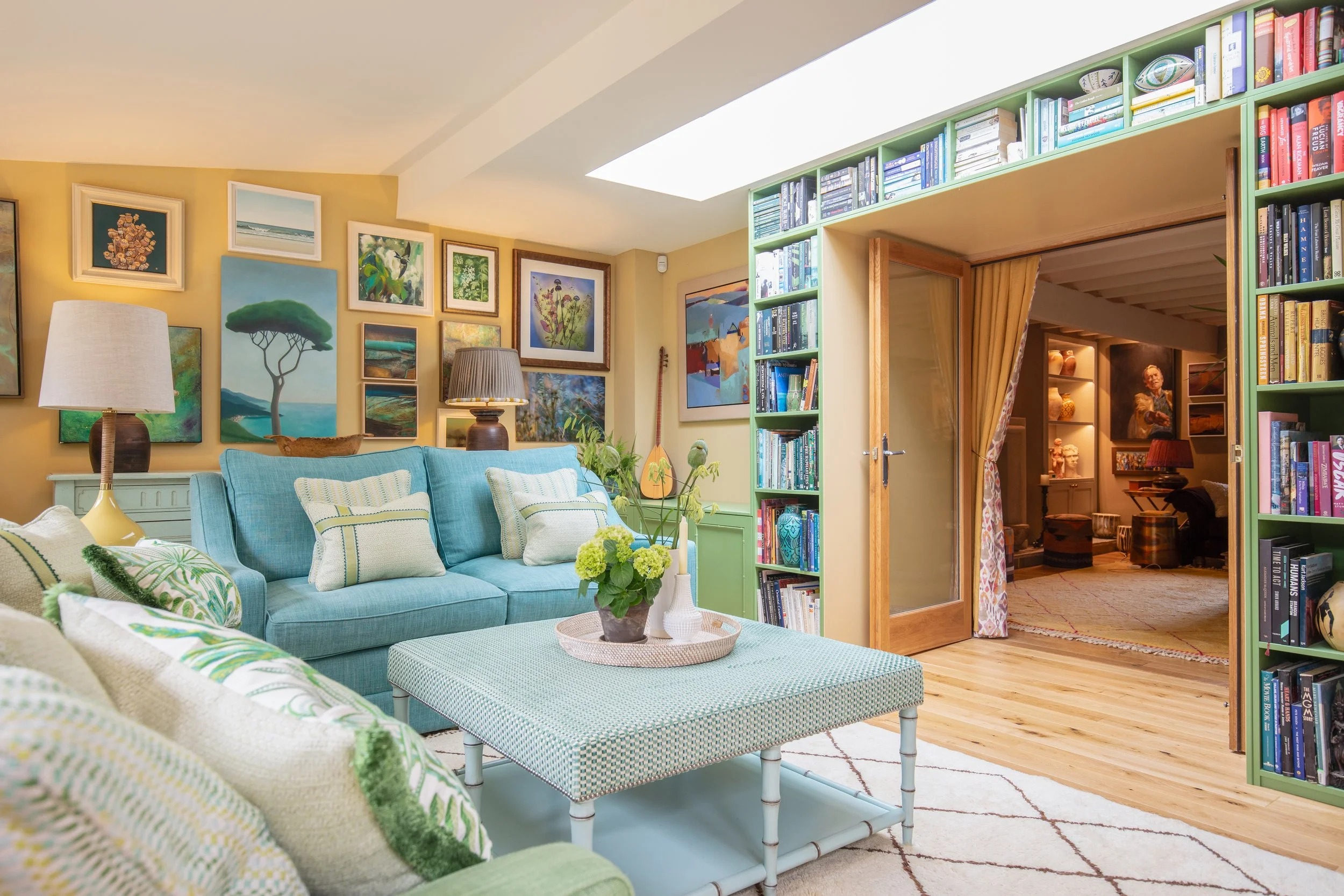 A cozy living room with a blue couch, green and white patterned pillows, a glass coffee table with a plant, and a wall decorated with colorful artwork. There is a green bookshelf filled with books and a sliding glass door with floral curtains leading