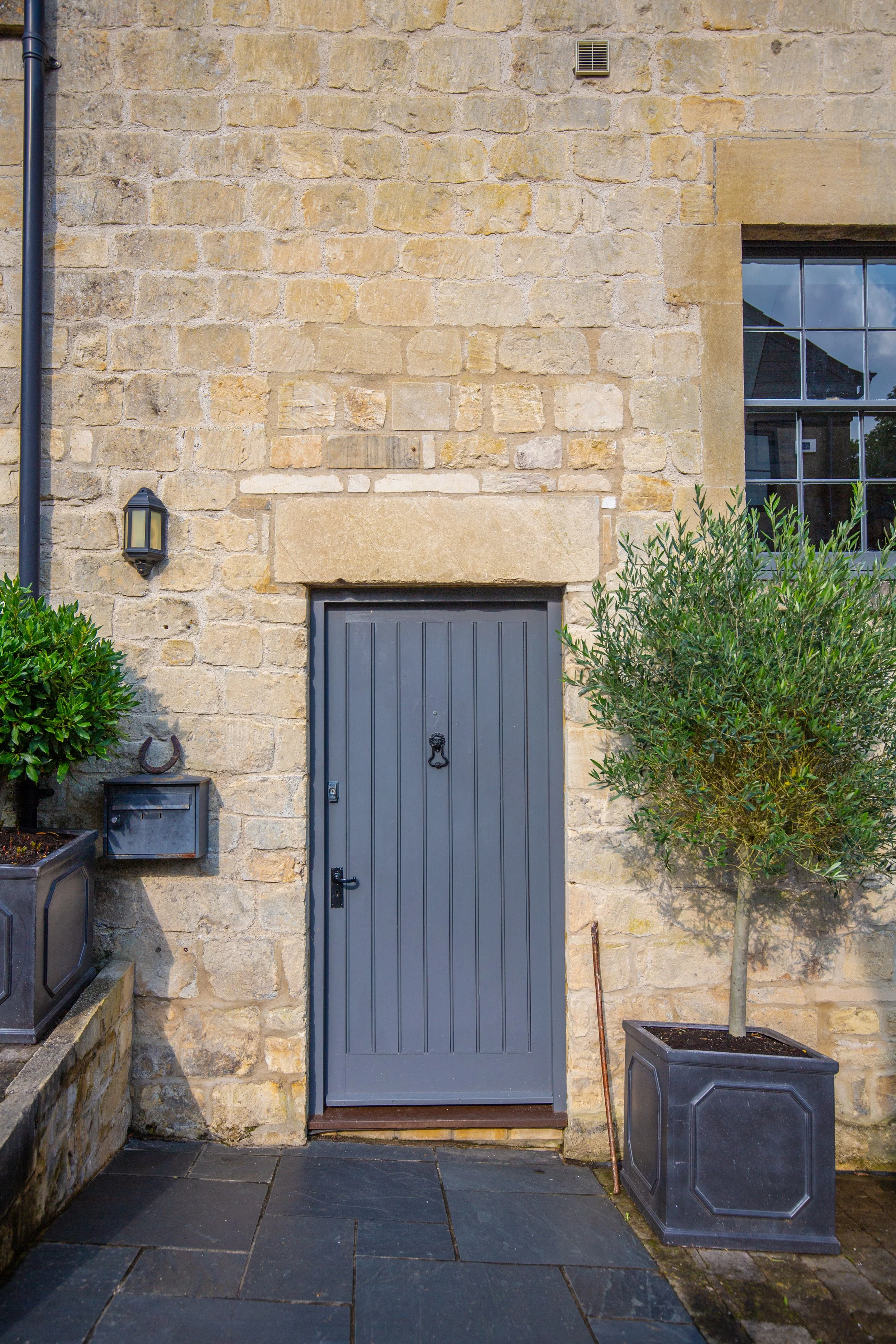 A stone wall with a small window on the right side and a dark gray door below it. There are two gray planters with green bushes on either side of the door. A black lantern-style light fixture on the left side of the door, a mailbox below it, and a ho