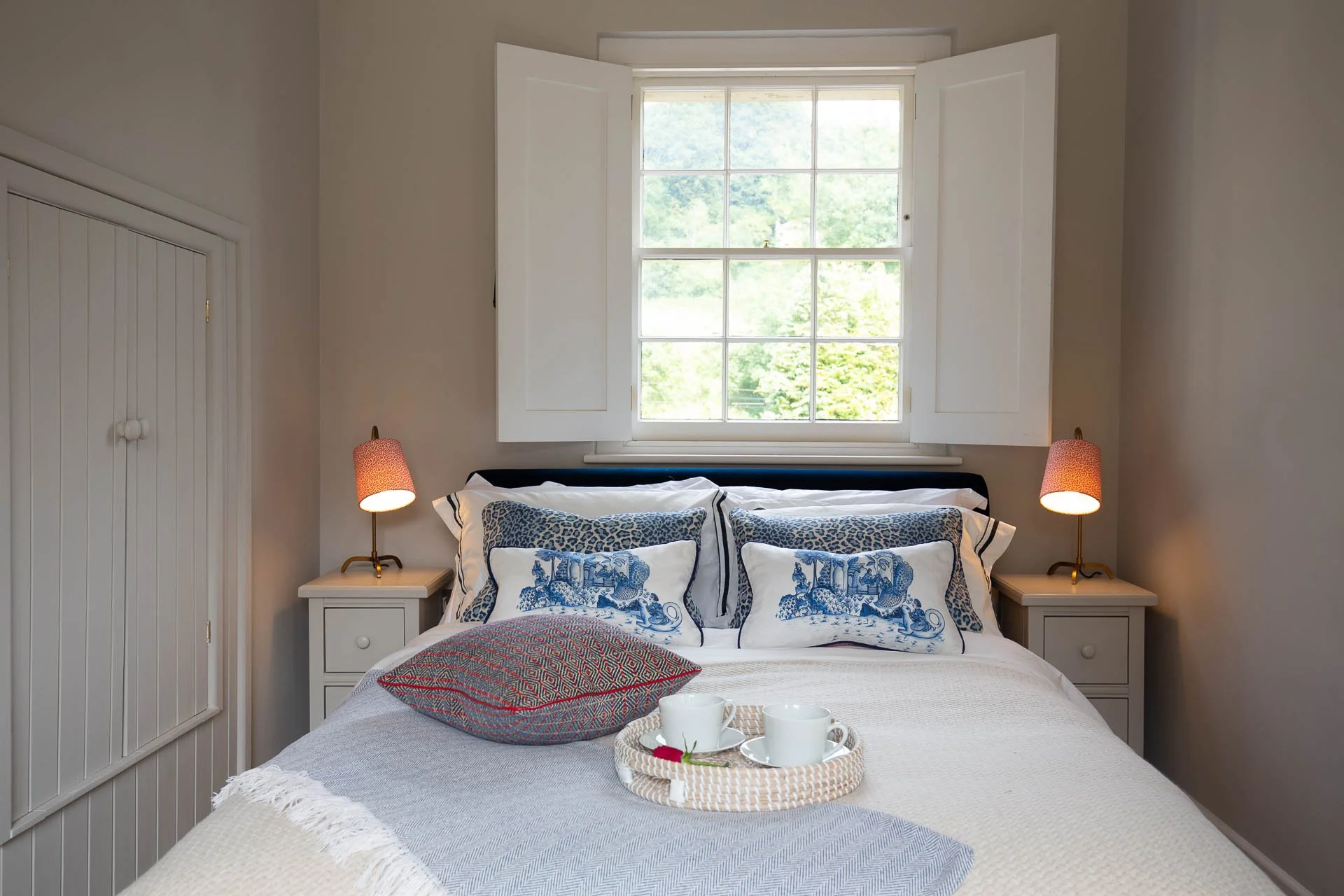 A cozy bedroom with a white bed, patterned pillows, tray with cups and roses, two bedside lamps, a window with open shutters, and beige walls.