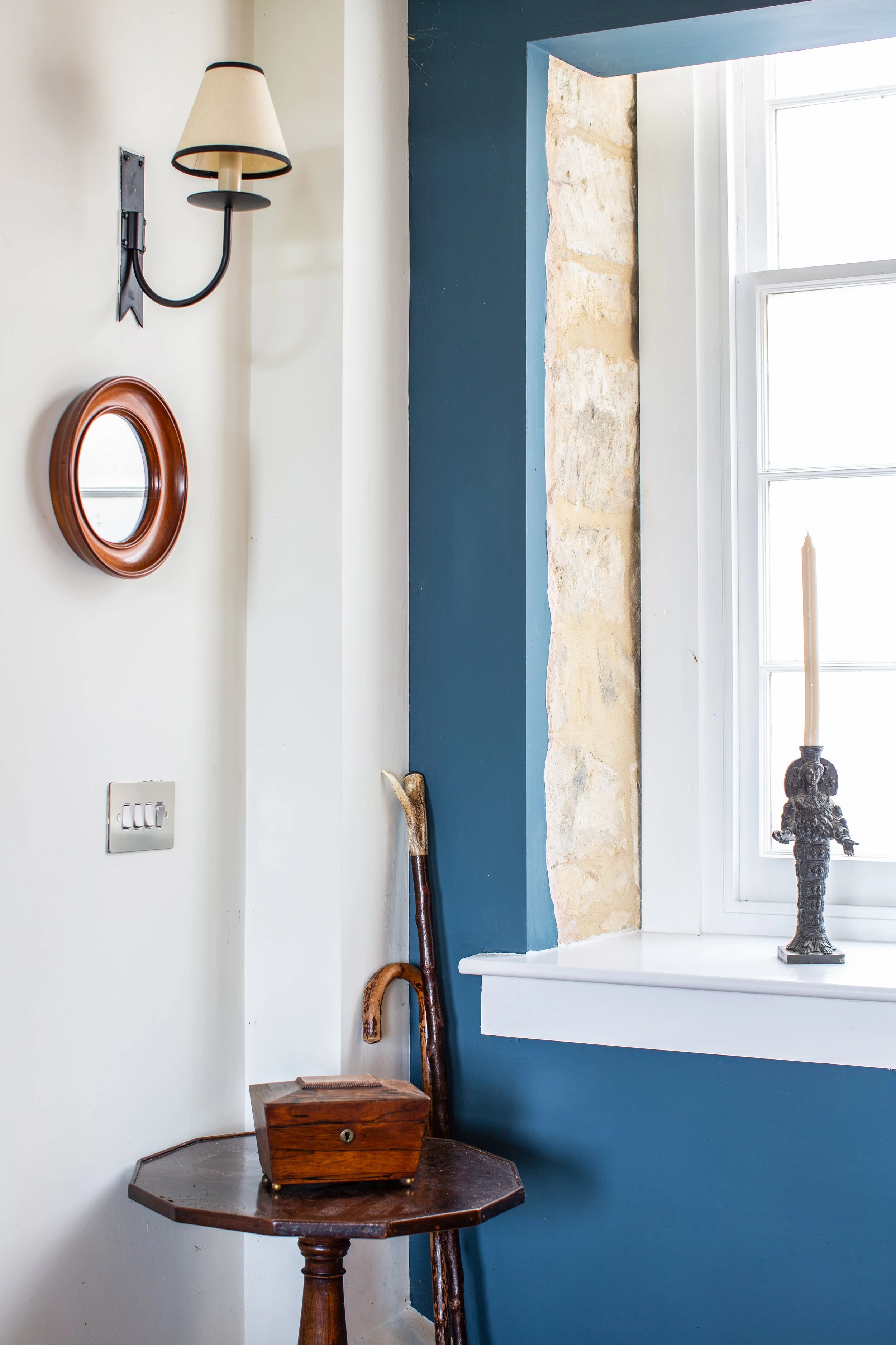 A corner of a room with a nightstand, a walking cane, a wall-mounted light fixture, a small round mirror, a window with a candle holder, and a bronze sculpture of a figure.