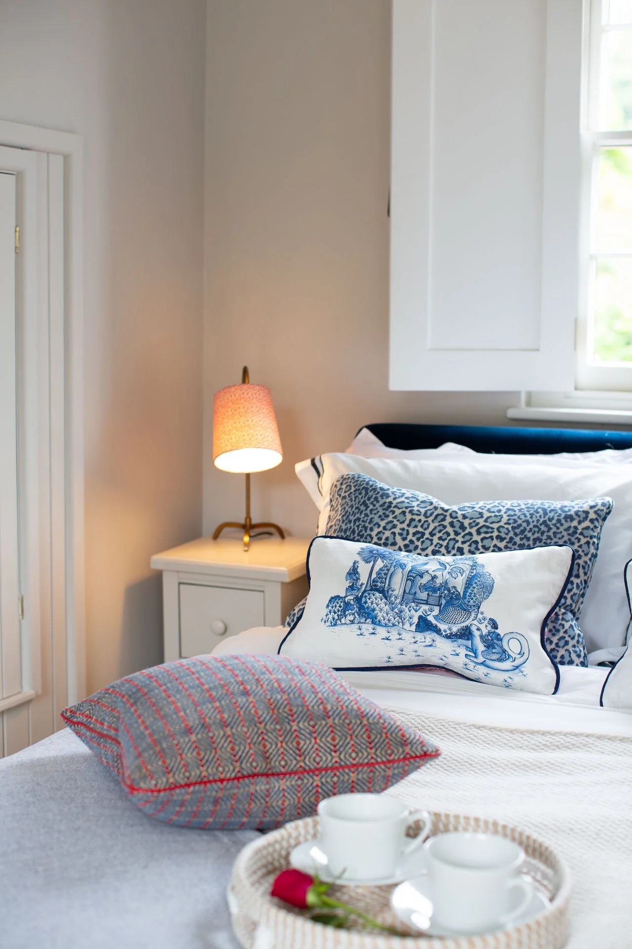 A cozy bedroom with a bed decorated with blue and white pillows, a white nightstand with a pink lampshade, a window with white shutters, and a tray with white cups and a pink flower in the foreground.