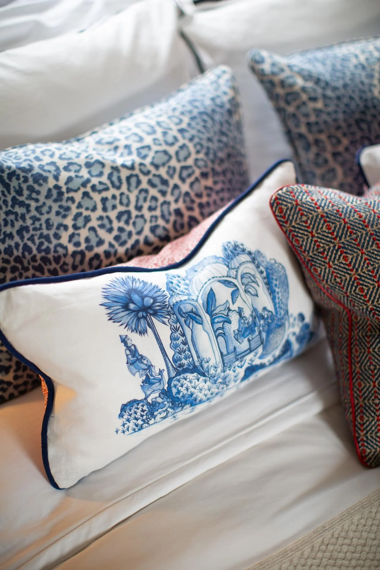 Close-up of decorative pillows on a bed featuring various patterns, including a blue and white toile, leopard print, and a geometric design with red accents.