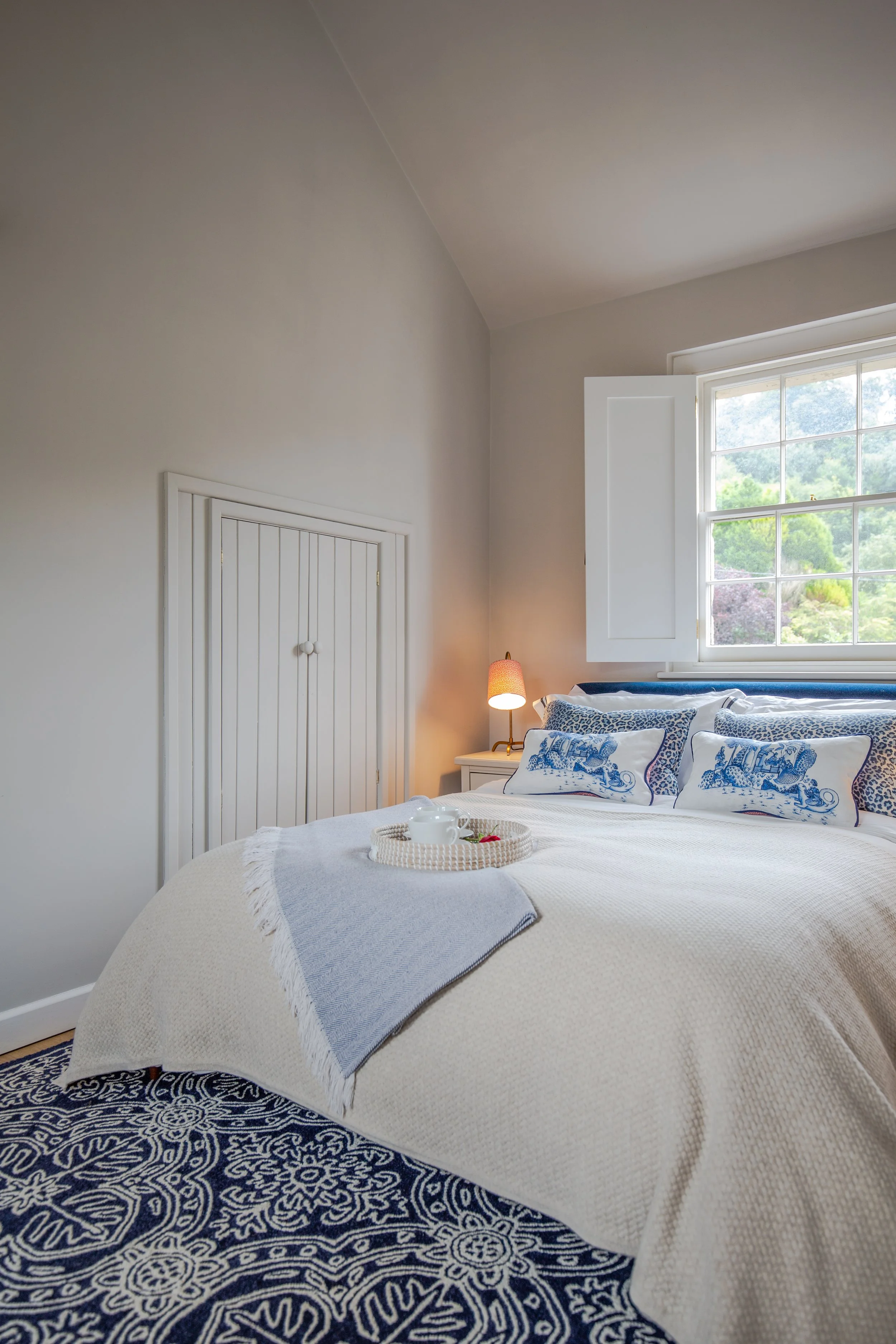 A cozy bedroom with a king-size bed covered in a white quilt, decorated with blue and white patterned pillows. A tray with a cup and strawberries rests on the bed. There is a window open, revealing green trees and mountains outside. A small white lam