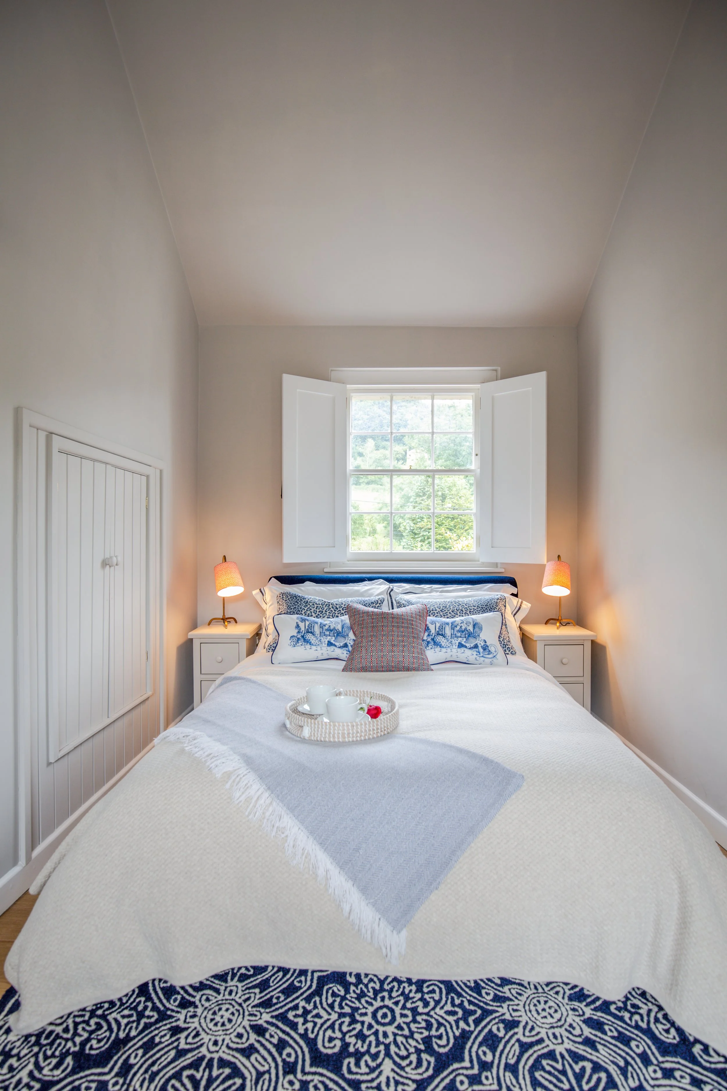 A neatly made bed with white bedding and a blue and white patterned rug underneath. There are two white nightstands with small lamps on each side of the bed. On the bed, there's a gray throw blanket and multiple pillows, including a decorative pillow