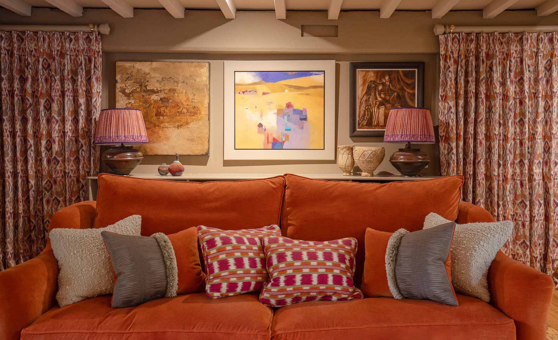 A living room with an orange sofa, patterned pillows, and a backdrop of three framed paintings on the wall, flanked by curtains with a multicolored geometric pattern and two table lamps.