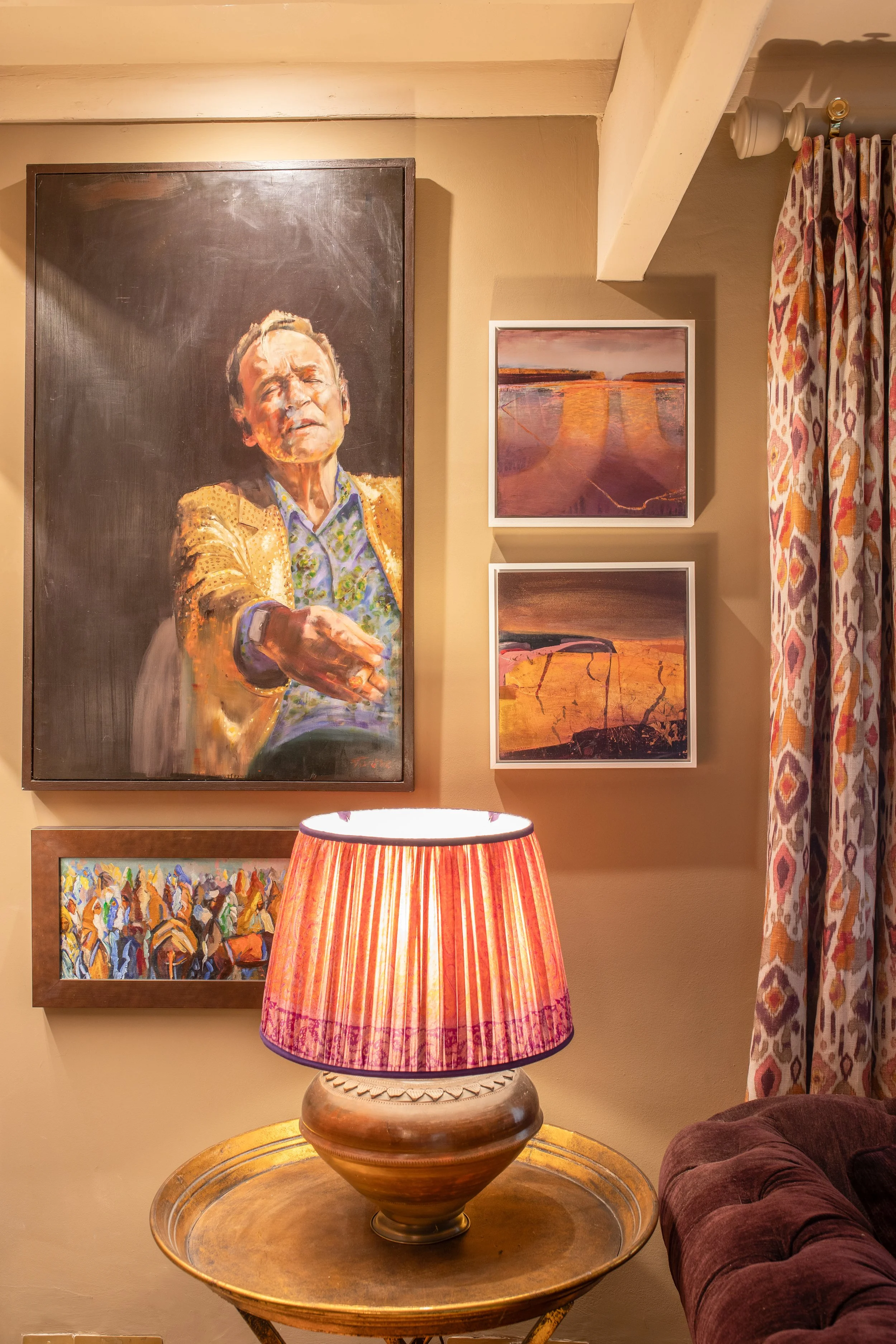 An interior scene featuring multiple framed paintings on a beige wall, including a large portrait of a man pointing, and a table with a warm-colored table lamp with a striped shade, next to a patterned curtain and a plush armchair.