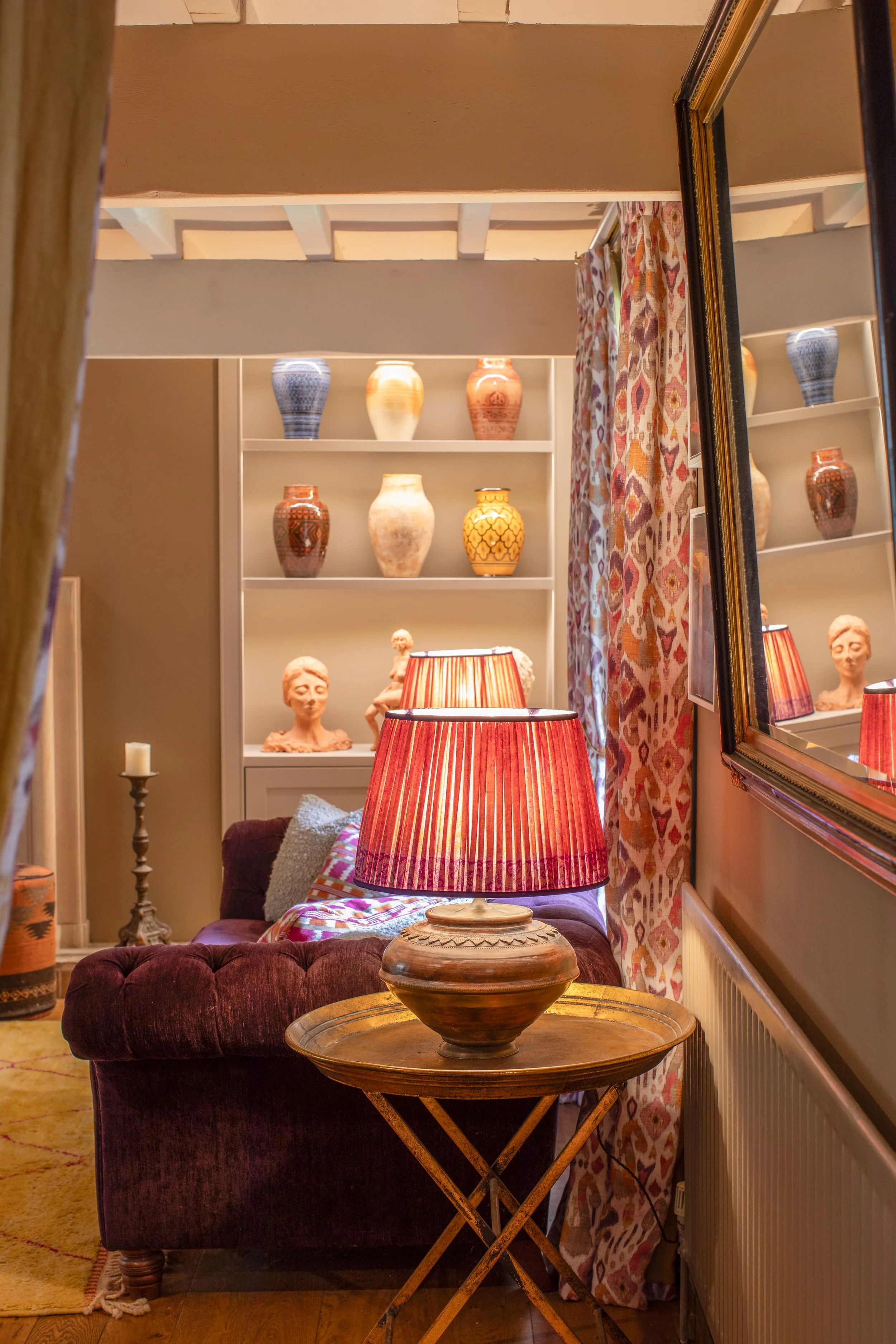A cozy living room corner featuring a deep red velvet armchair with a pillow, a small wooden side table holding a decorative bowl, a table lamp with a red and orange striped shade, a white bookcase with ceramic vases and bust sculptures, and patterne