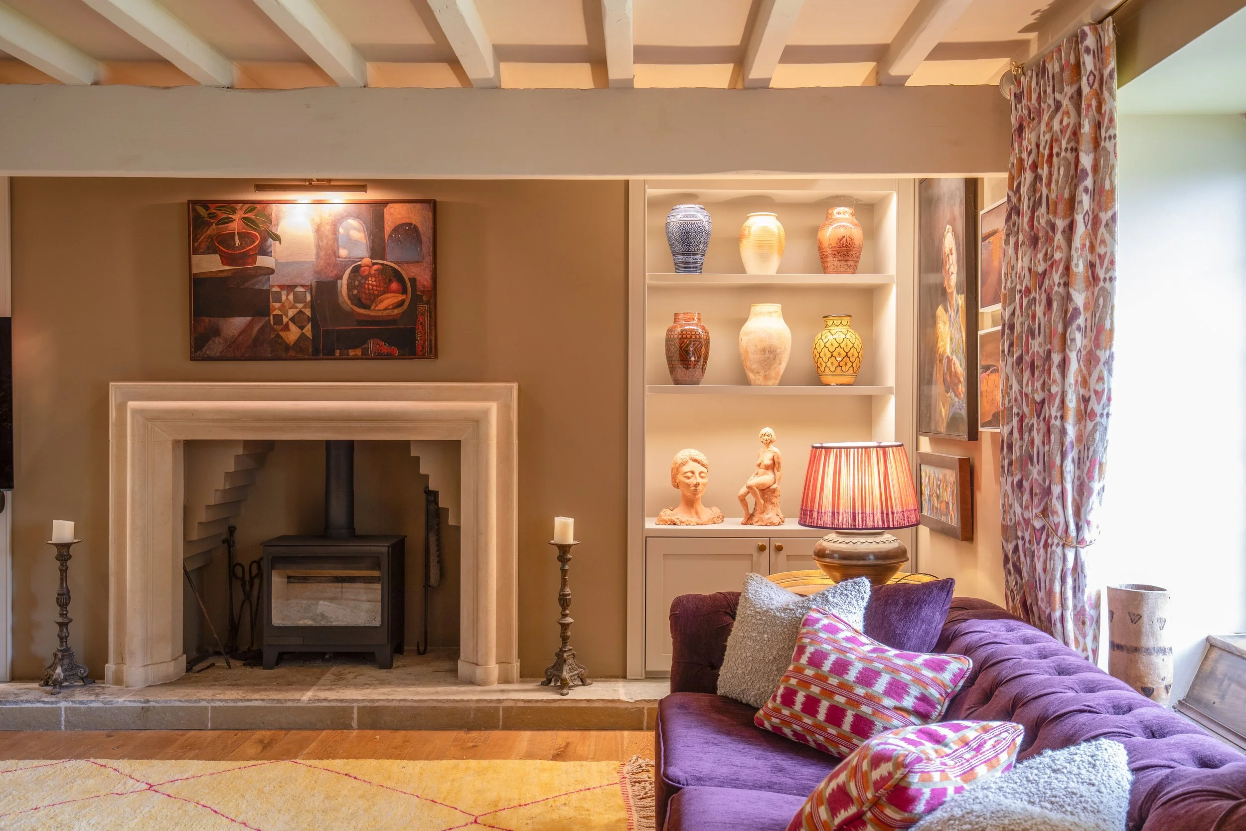 Living room with a purple velvet sofa and colorful patterned cushions, a wood-burning stove in a fireplace with white marble trim, a bookshelf with decorative vases, a framed painting above the fireplace, and a window with floral curtains.