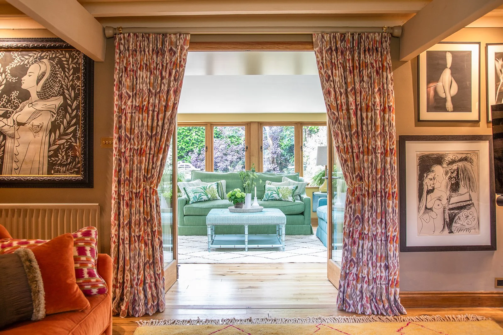 Living room looking out onto a sunroom with green sofa and large windows, framed artworks on the walls, patterned curtains, and a cozy, colorful atmosphere.
