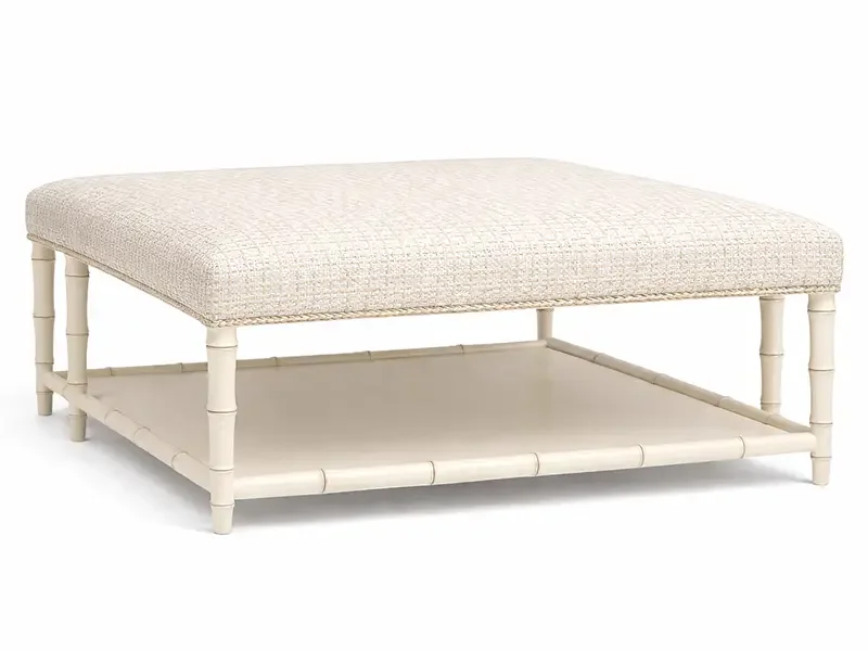 A beige upholstered ottoman with a lower shelf and bamboo-style legs.