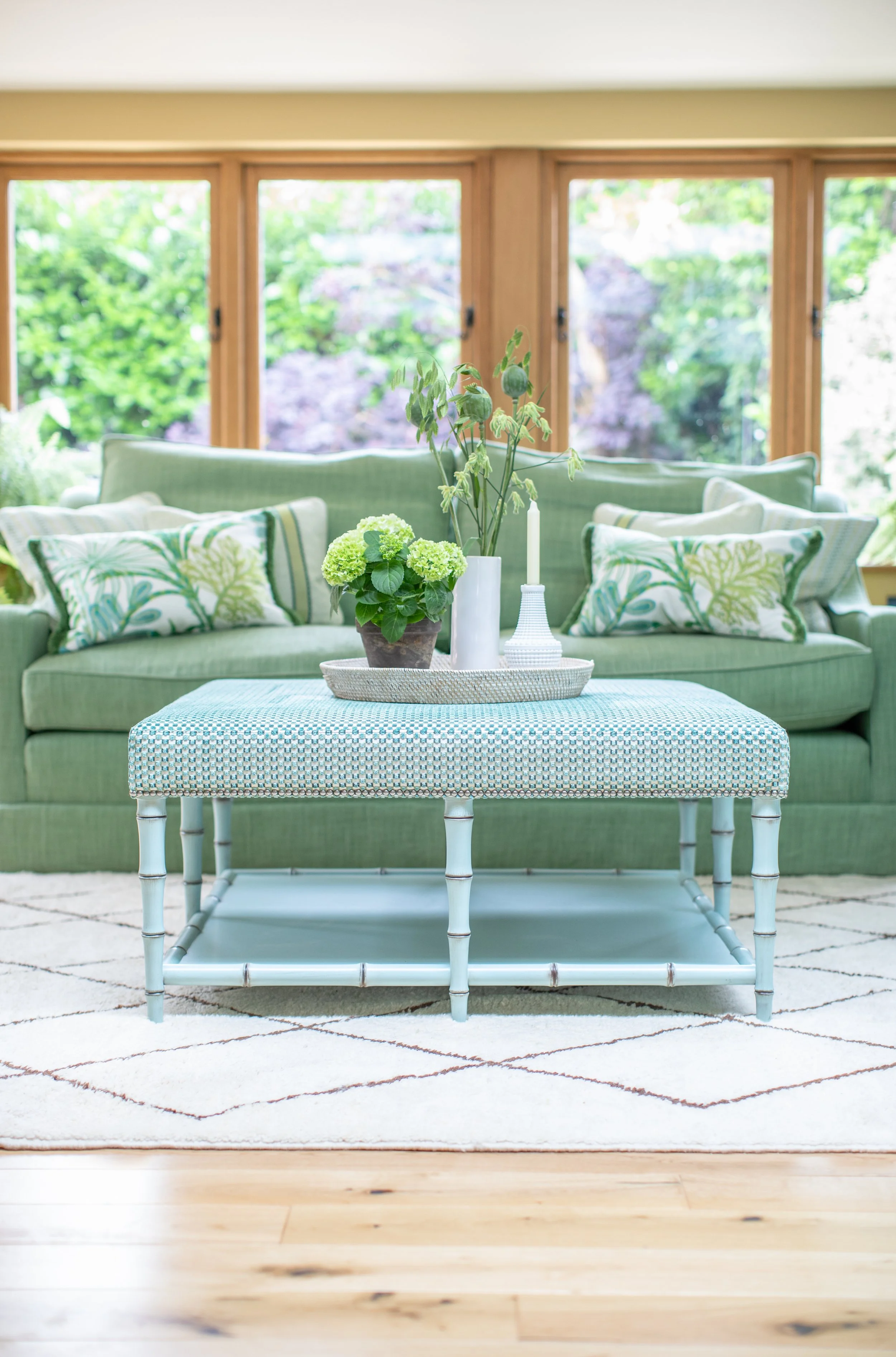 A living room with a green sofa, patterned pillows, a wicker coffee table with plants, and large windows showing greenery outside.