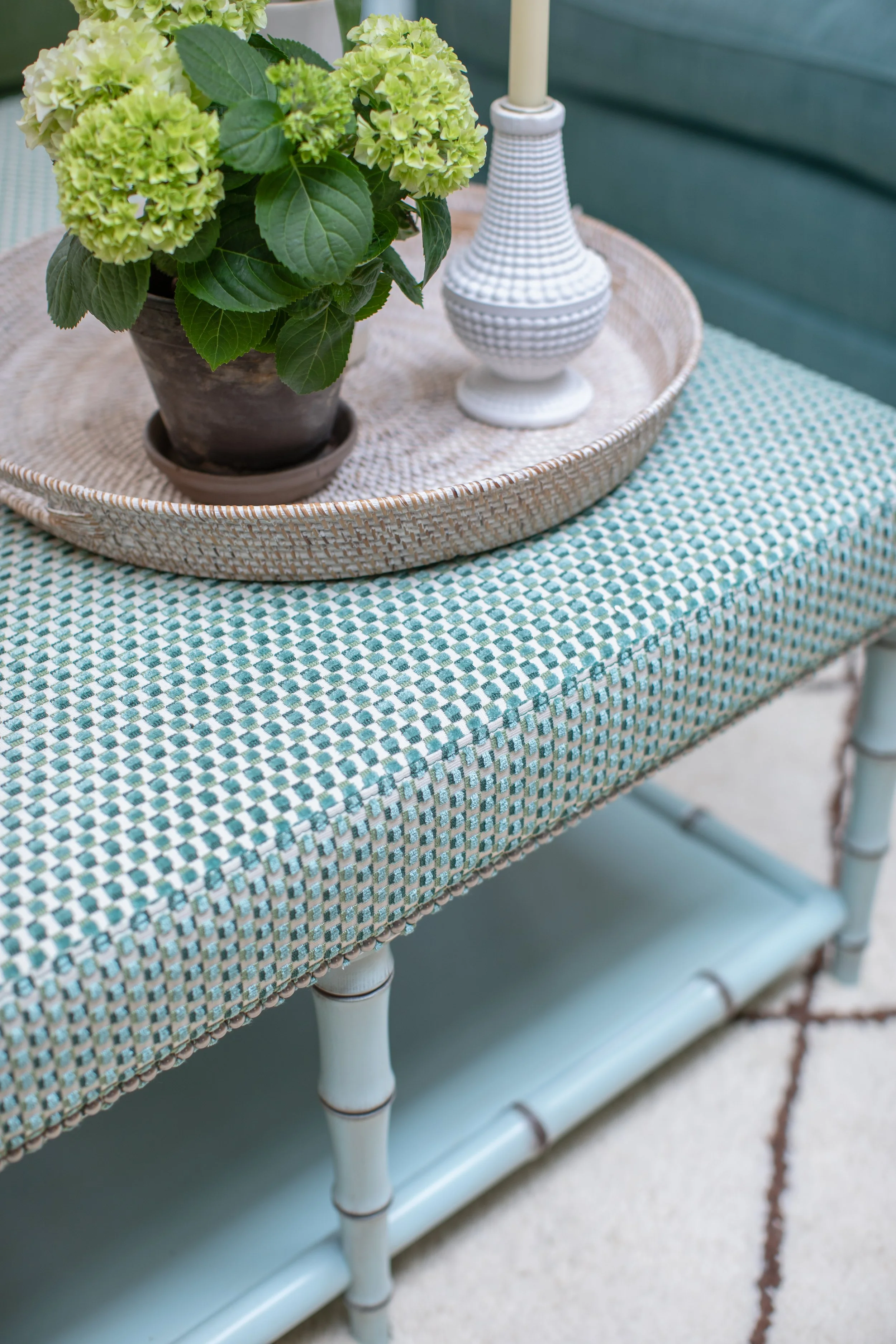 A close-up view of a table with a blue and white checkered tablecloth, a woven wicker tray with a potted green hydrangea plant, and a white ceramic cottage-style lamp with a cream-colored lampshade. The background includes a blue wooden wall.