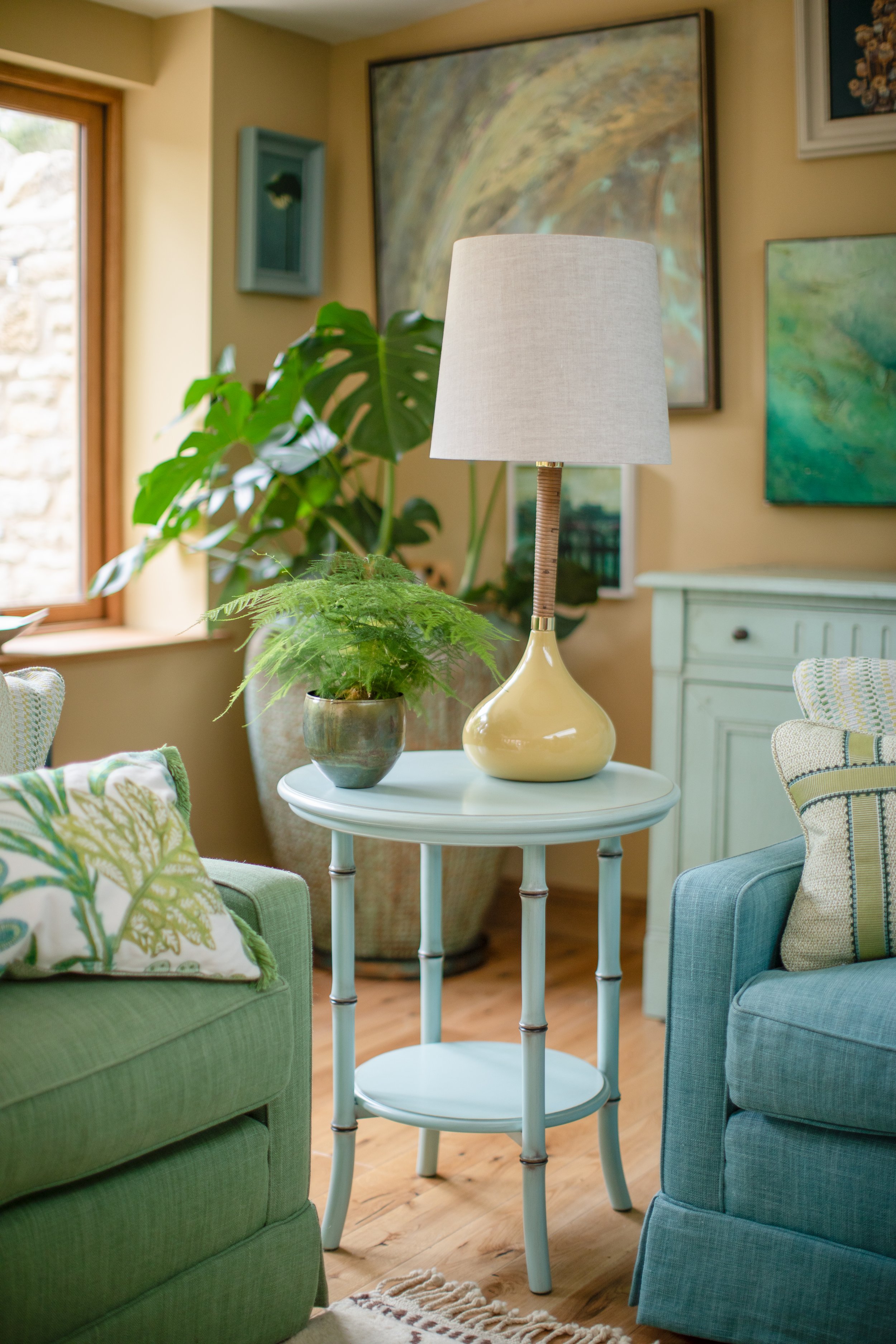 Living room with green and blue armchairs, a white side table with a lamp, and artwork on the walls. There's a large window with natural light.