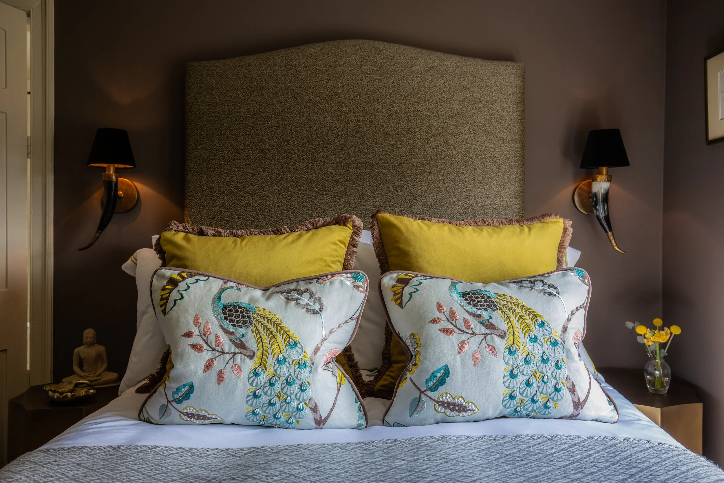 A neatly made bed with two decorative pillows featuring embroidered floral and bird designs, and a textured blanket. Two black wall-mounted lamps with gold accents flank a large, padded headboard against a dark-colored wall, with a small Buddha statu