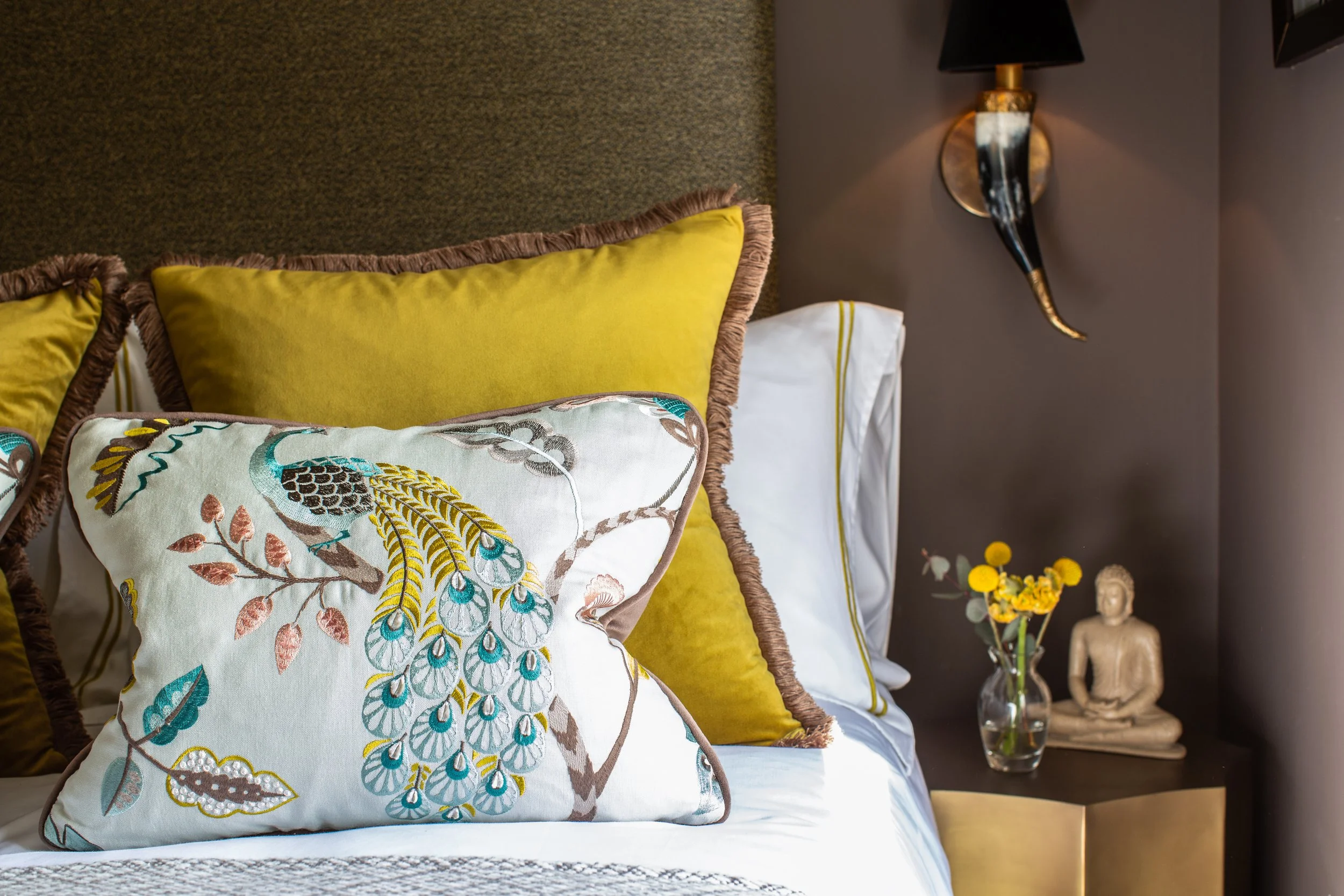 A bedroom with yellow and decorative pillows on a bed, a wall-mounted lamp, a side table with a glass of water, a small statue, and a bouquet of yellow flowers.