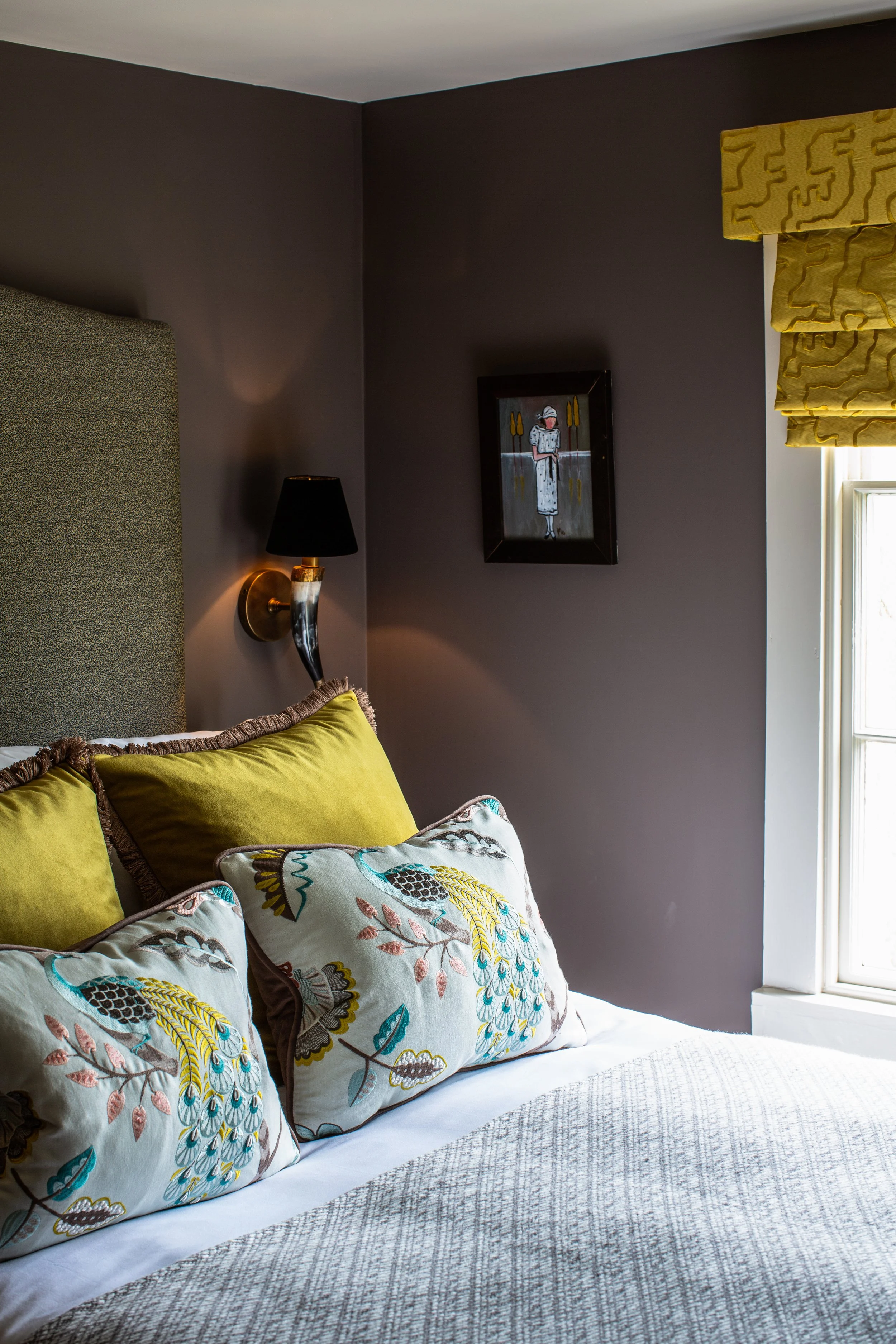A bed with decorative pillows, a wall lamp, dark gray wall, framed artwork, and window with yellow patterned Roman shades in a bedroom.
