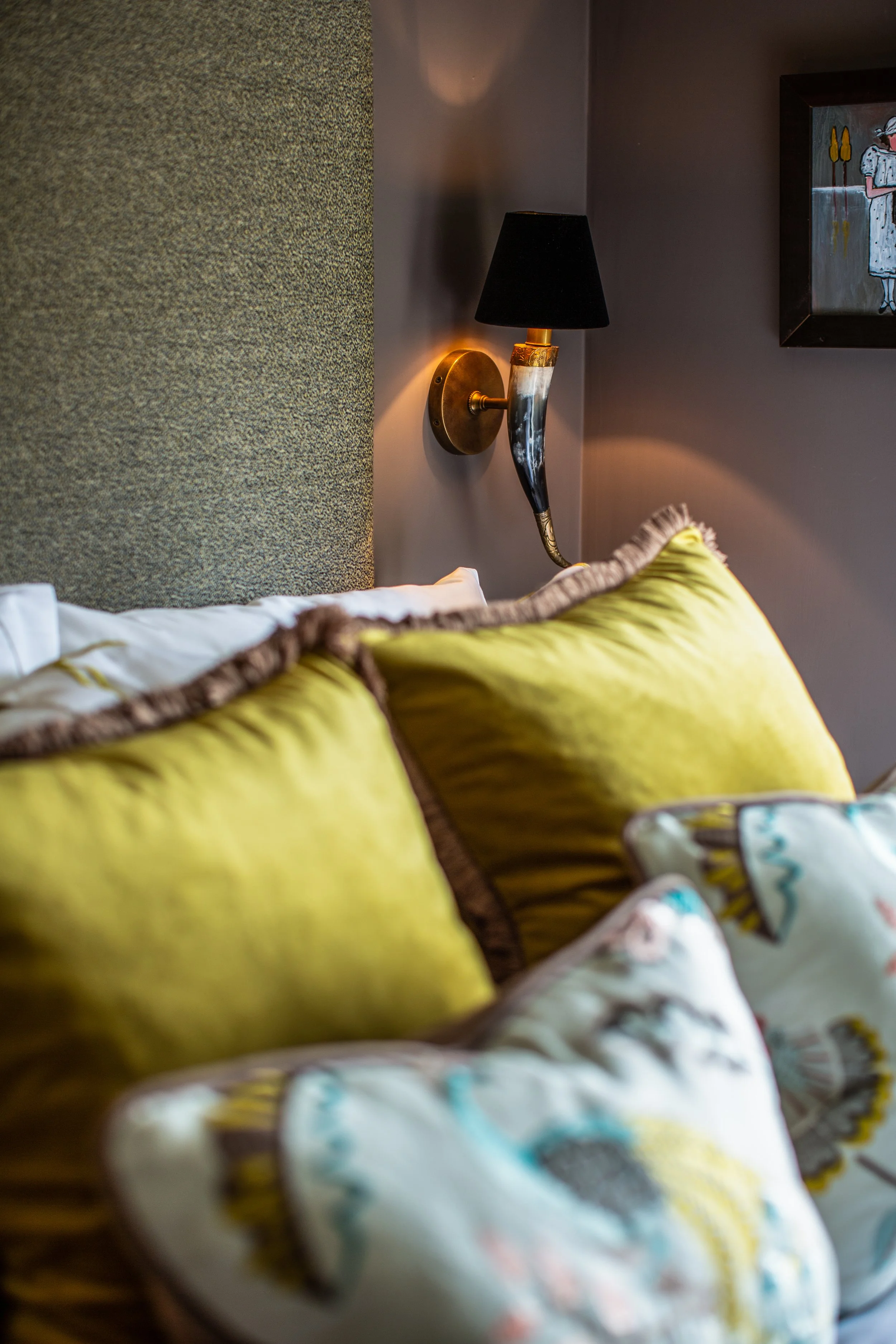 Close-up of pillows on a bed with a dark wall, framed artwork, and a wall-mounted lamp in the background.