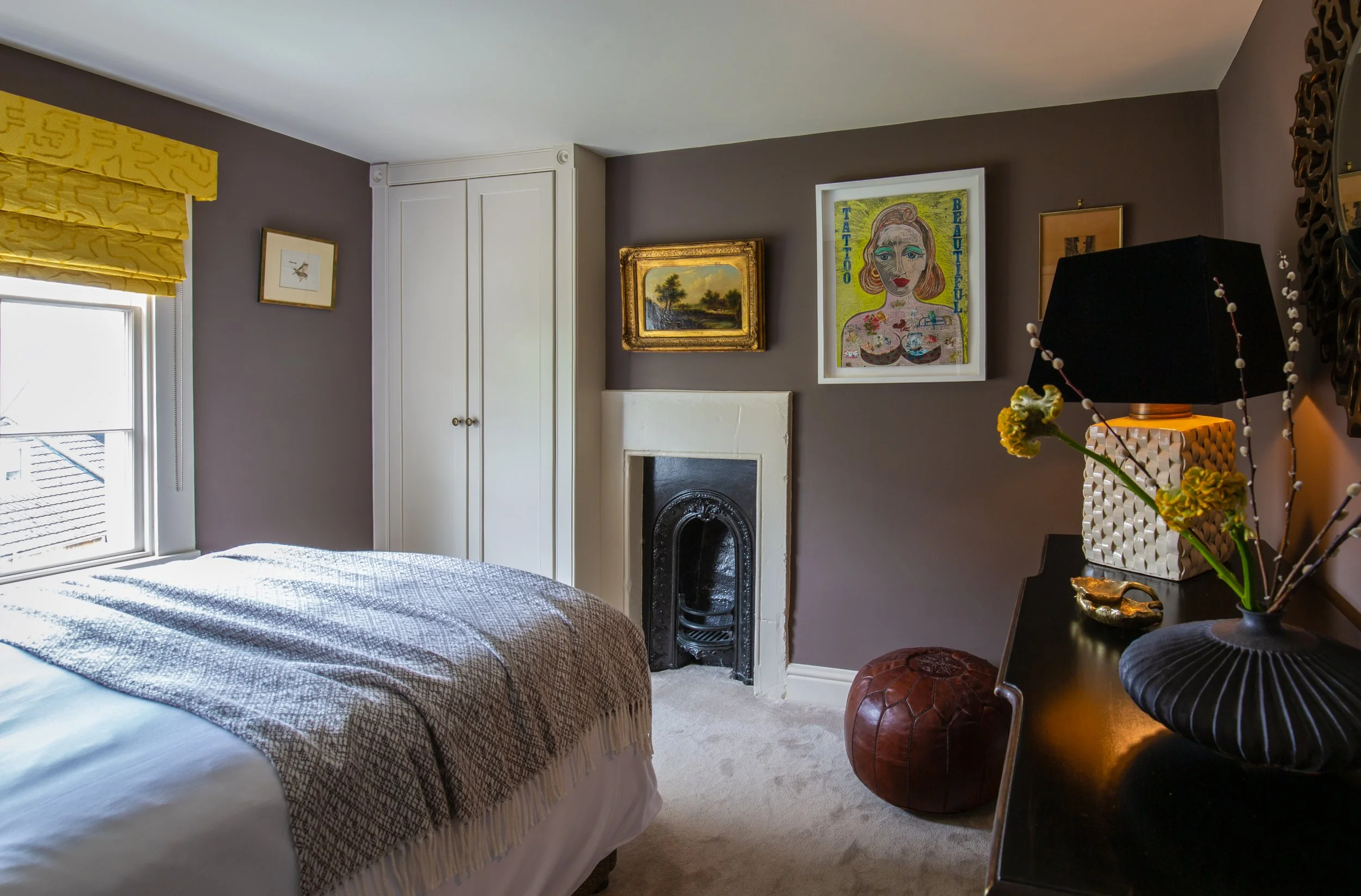A cozy bedroom with a window, decorated with yellow curtains, and a bed with a checkered blanket. The room features a white fireplace, framed artwork, and a side table with a black lamp, a yellow vase, and fresh flowers.