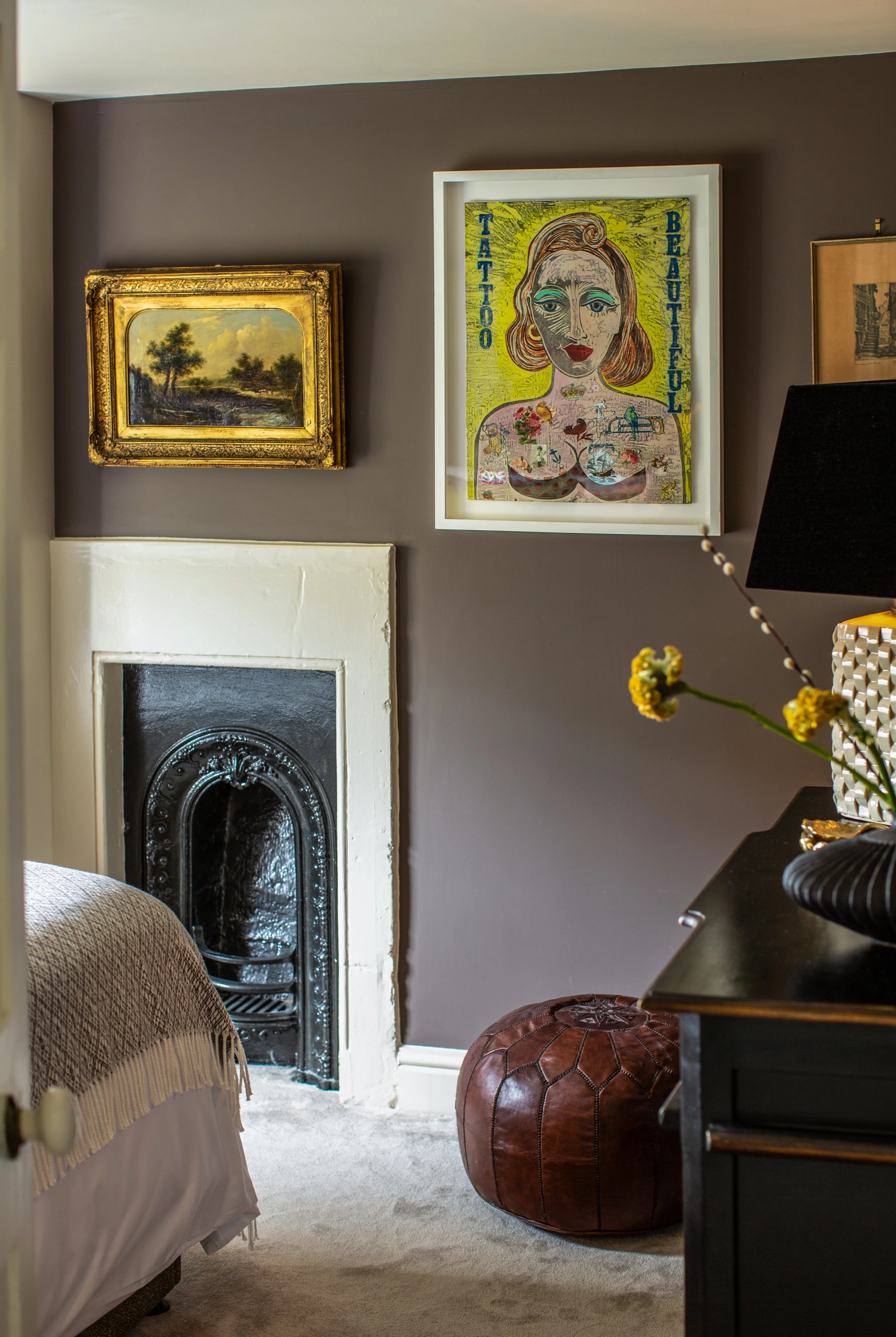 Living room with a fireplace, framed artwork on the wall, a leather pouf, a black table with decorative objects, and a flower vase on the right side.