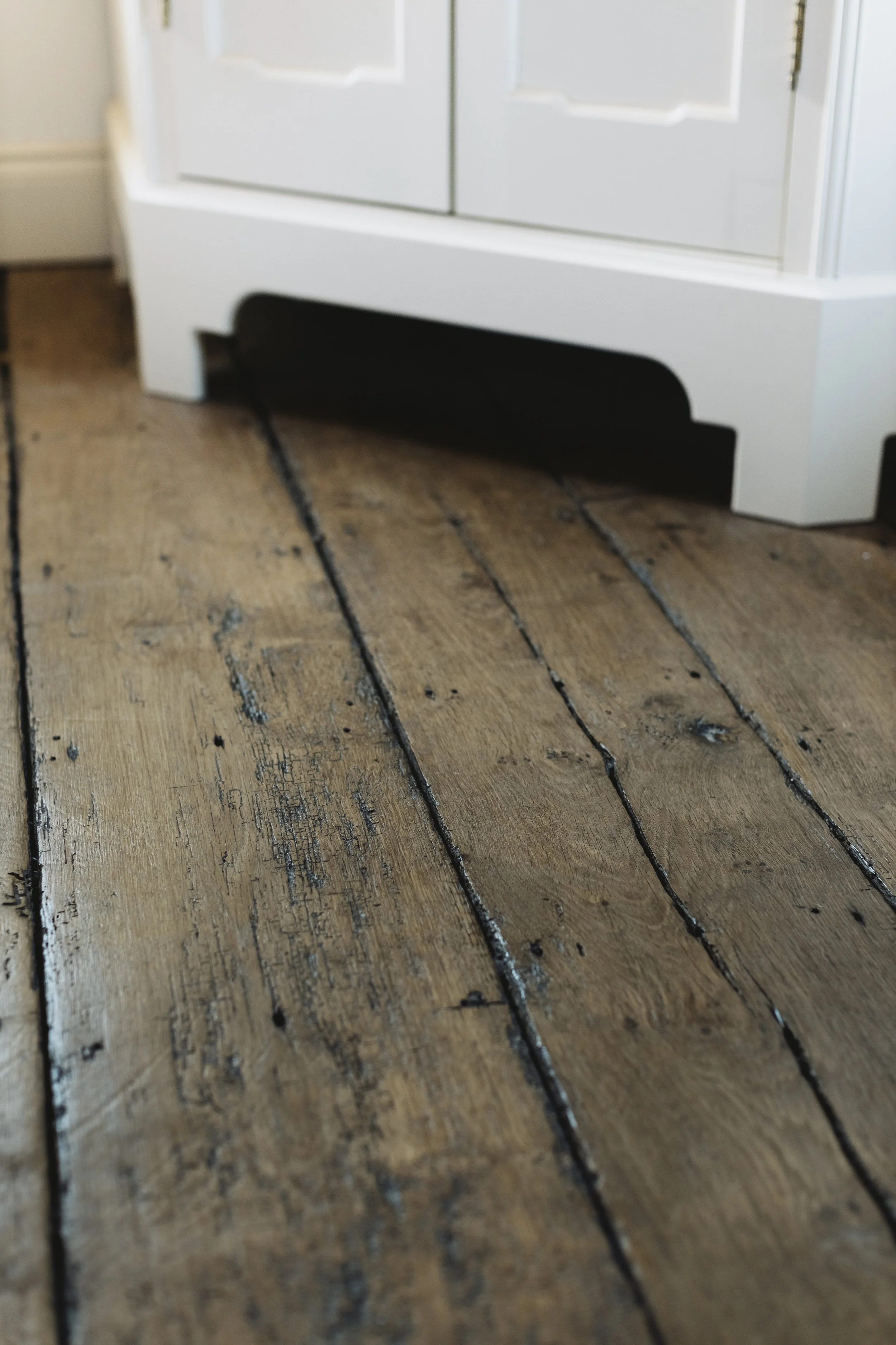Close-up of a wooden floor with visible grain and blackened nails, with a white furniture piece partially visible at the top.