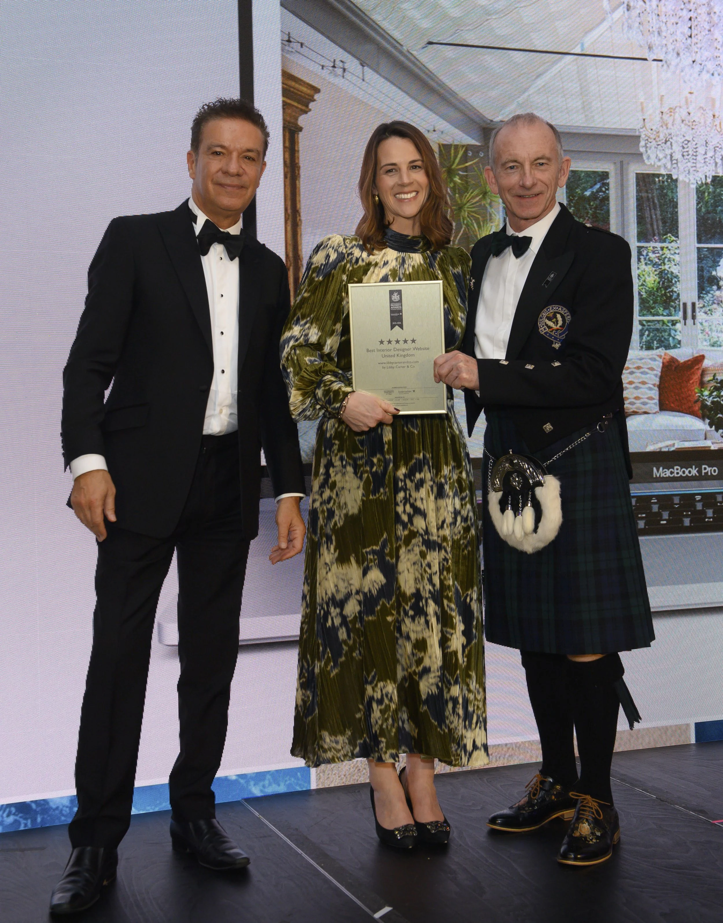 Three individuals standing on a stage, two men in tuxedos and a woman in a patterned dress, holding an award plaque.