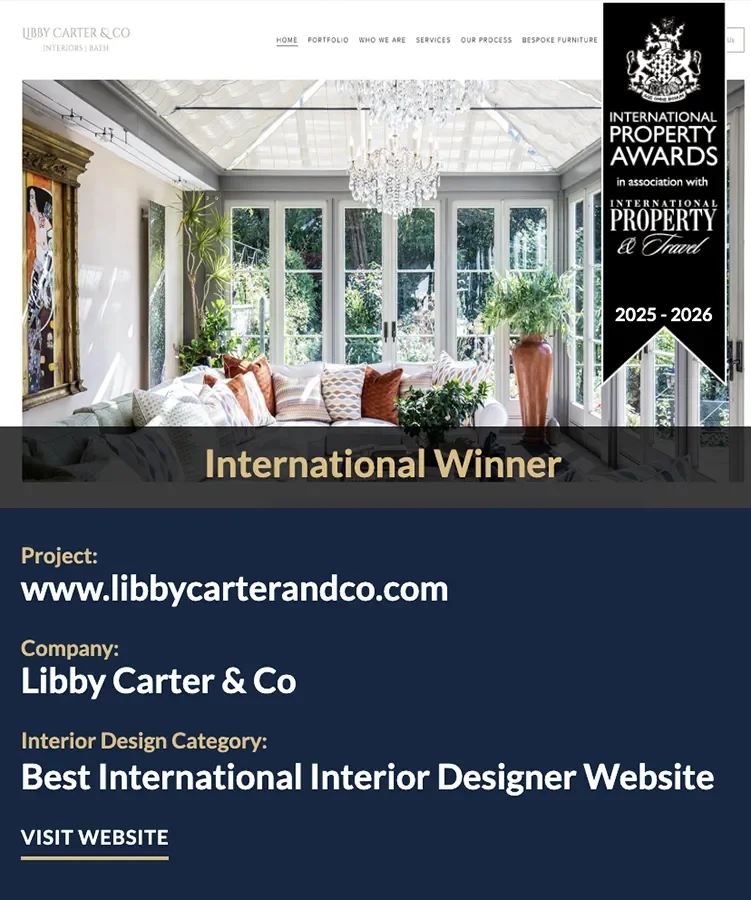 An award announcement for Libby Carter & Co, an interior design company, featuring a bright, elegant room with large windows, a chandelier, and a white sofa with throw pillows. The banner on the right indicates the company won the 'Best International Interior Design Website' award for 2025-2026.