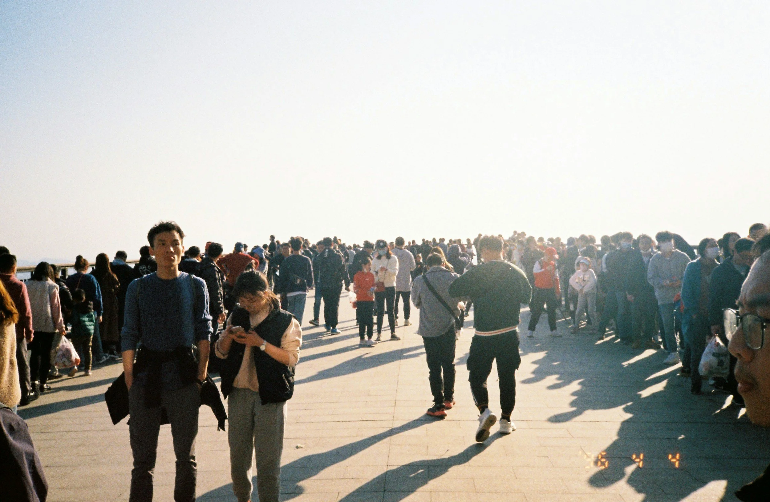 Crowd of people gathered outdoors on a sunny day, some wearing masks, some looking at their phones, with the sky and horizon in the background.