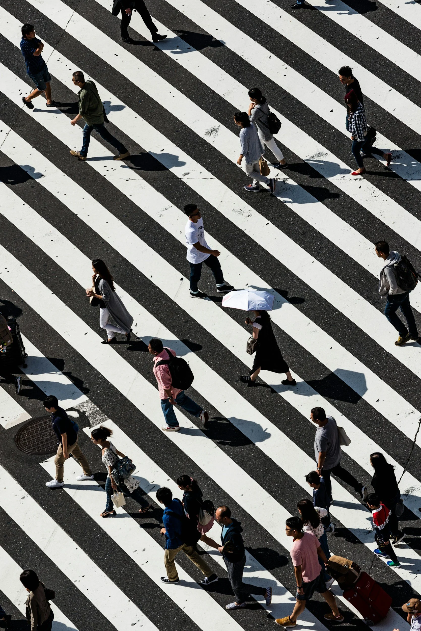 People walking across a crosswalk on a city street, casting shadows on the pavement.