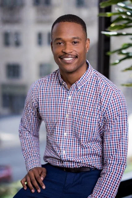 A man with short hair, a mustache, and a smile, wearing a checkered button-up shirt and dark pants, standing indoors near a window with a cityscape in the background.