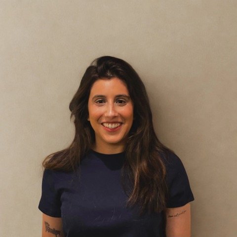 A woman with long dark hair and a big smile, wearing a dark blue shirt, standing against a plain beige wall.