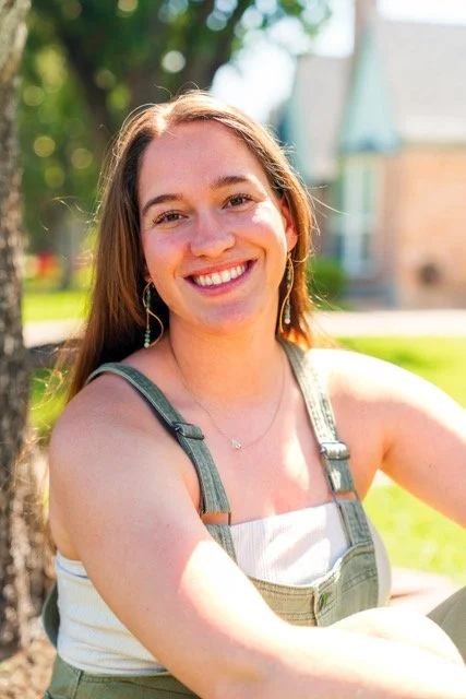 A young woman with long brown hair smiling outdoors on a sunny day, wearing a sleeveless top and overalls.