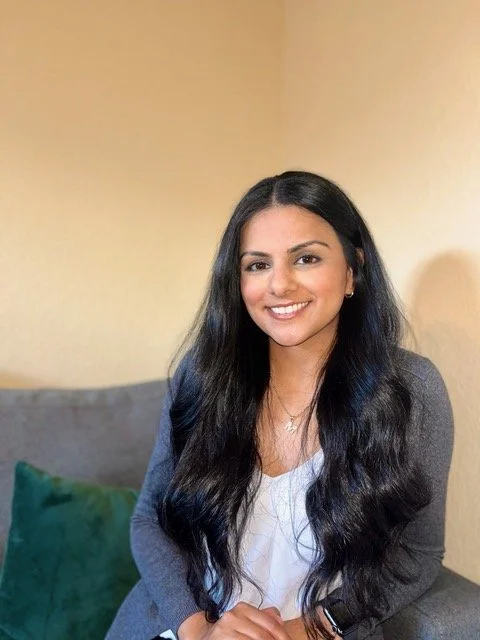 A woman with long, dark hair is smiling and sitting on a couch with a beige wall behind her.