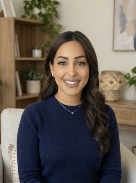 A smiling woman with dark brown hair and wearing a navy blue sweater, sitting indoors with a bookshelf, plants, and a painting in the background.