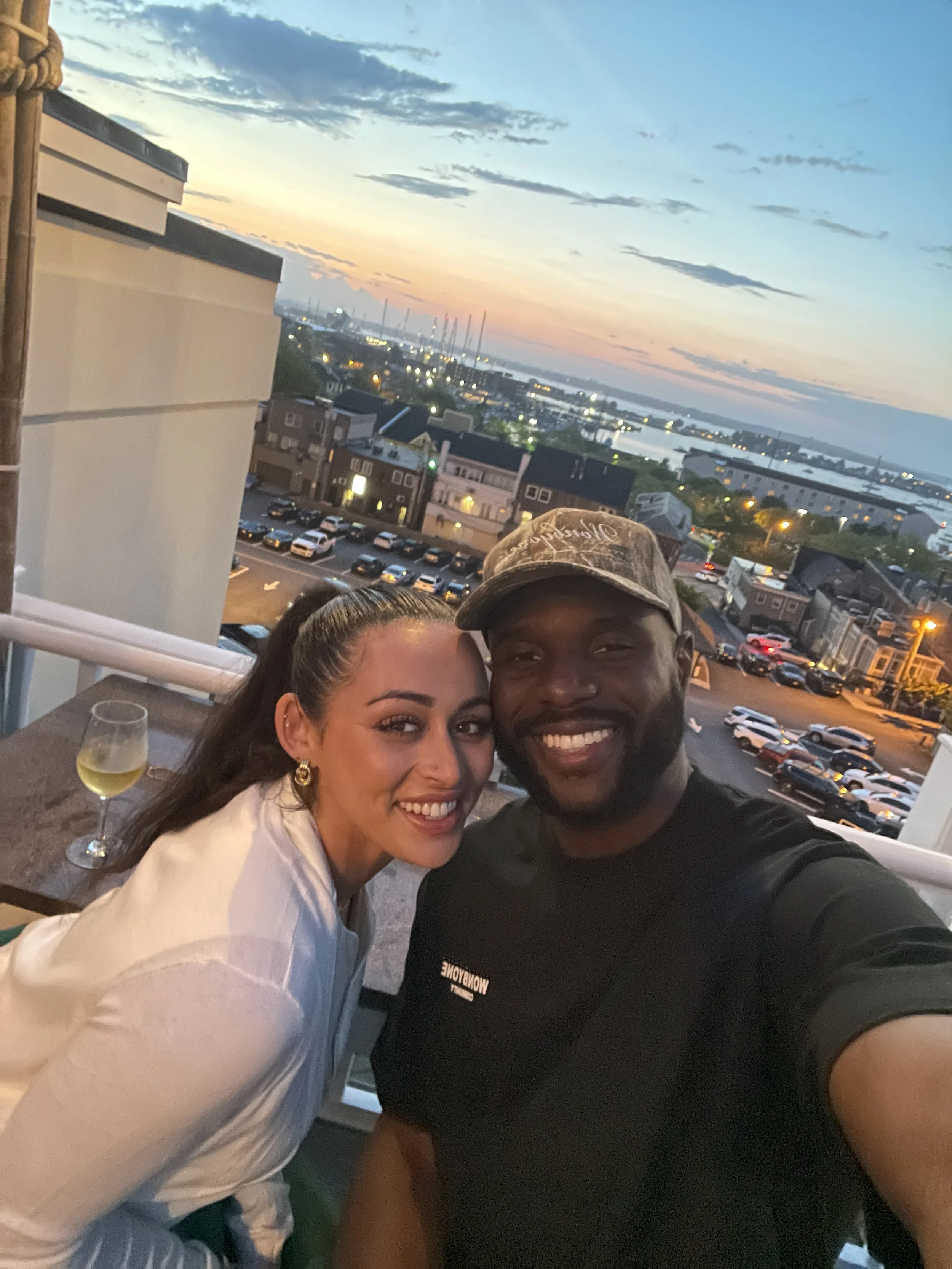 Smiling couple taking a selfie by a window overlooking a marina and waterfront city at sunset