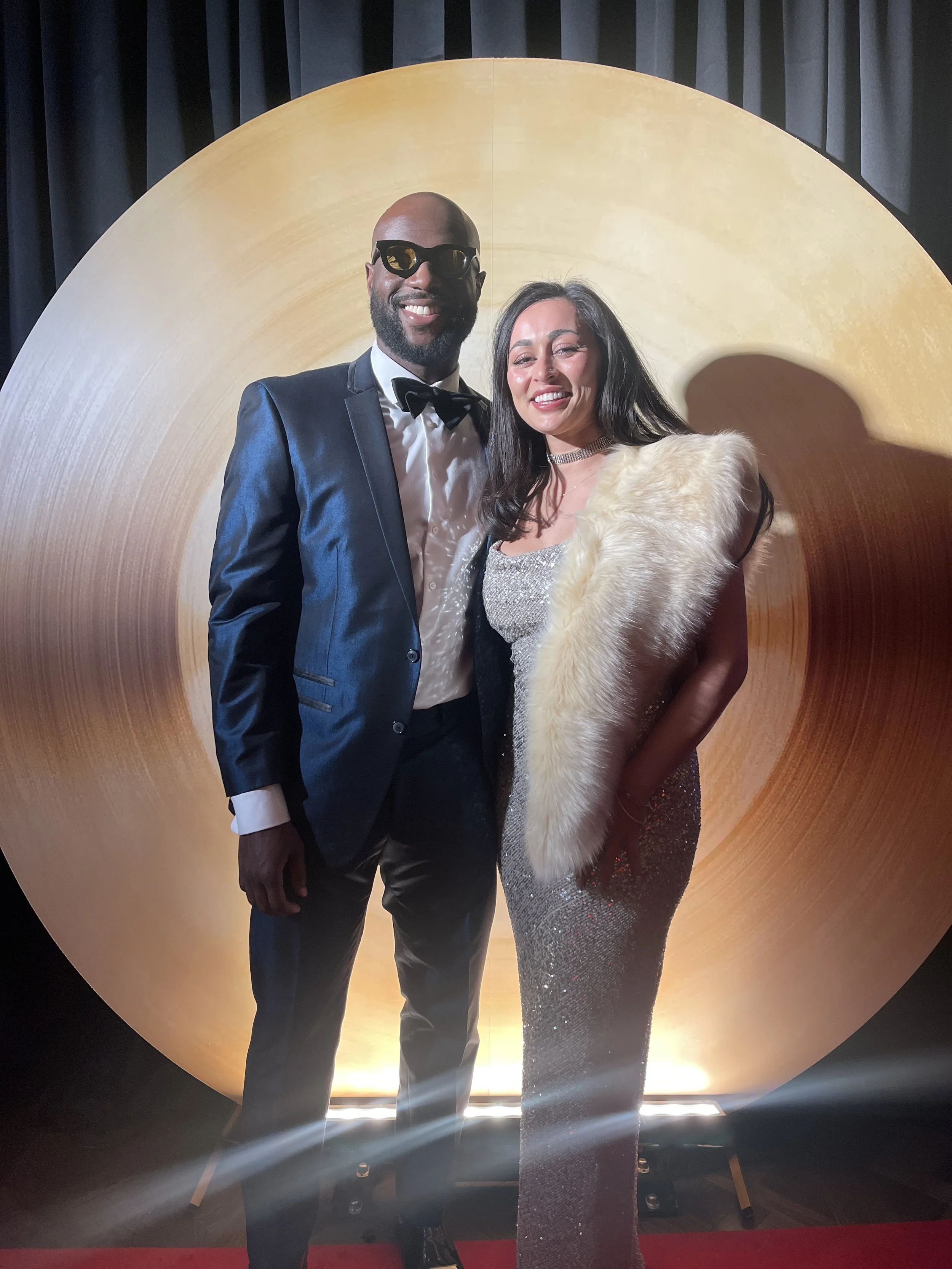 Smiling couple dressed in formal evening attire posing on a red carpet in front of a large golden circular backdrop