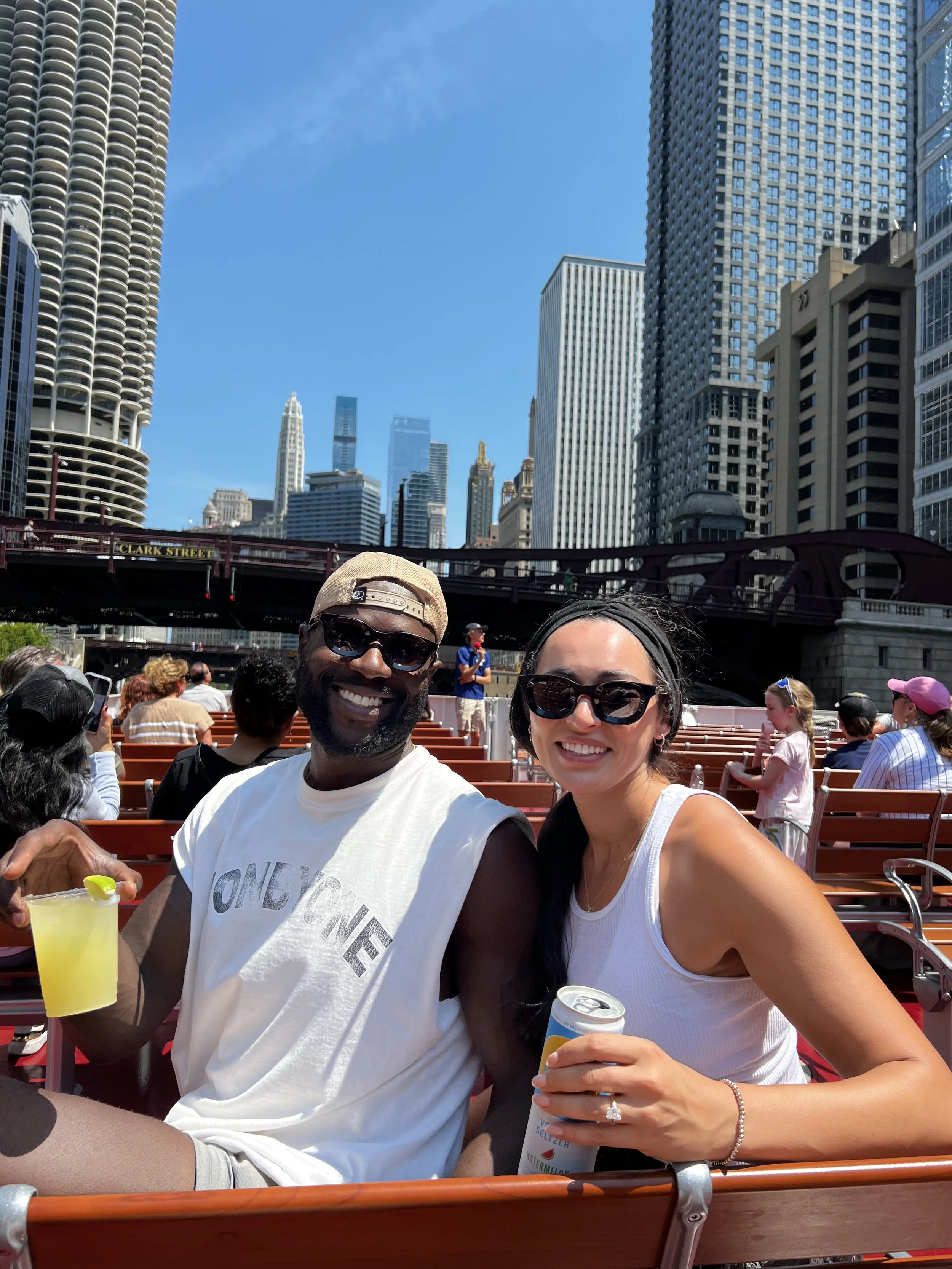 Couple taking a selfie on an open-air Chicago architecture boat tour with downtown skyscrapers and the Clark Street bridge behind them.