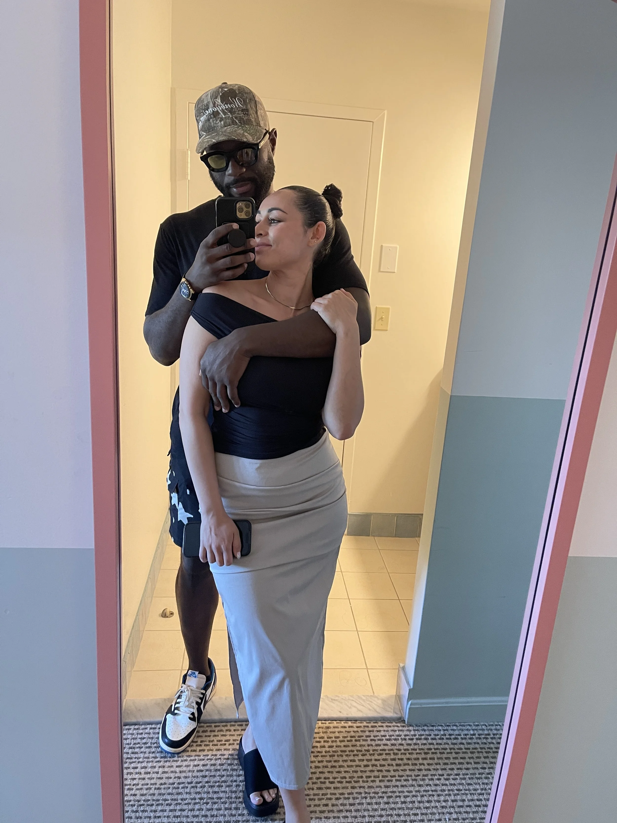 Couple taking a mirror selfie, with a man wearing a camouflage cap and sunglasses standing behind a woman in a black top and gray skirt.