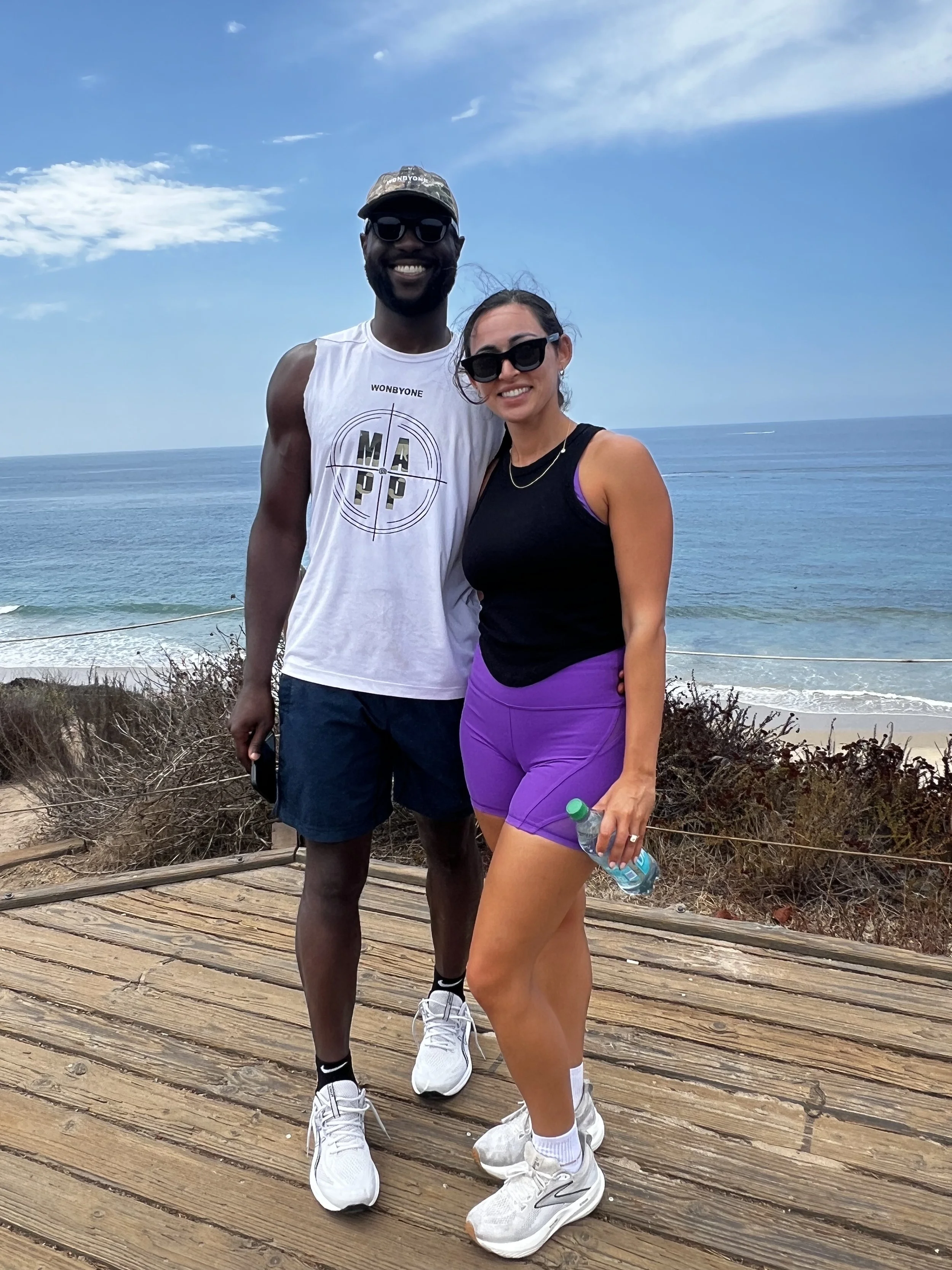 Smiling couple wearing sunglasses poses on a wooden boardwalk overlooking the ocean.