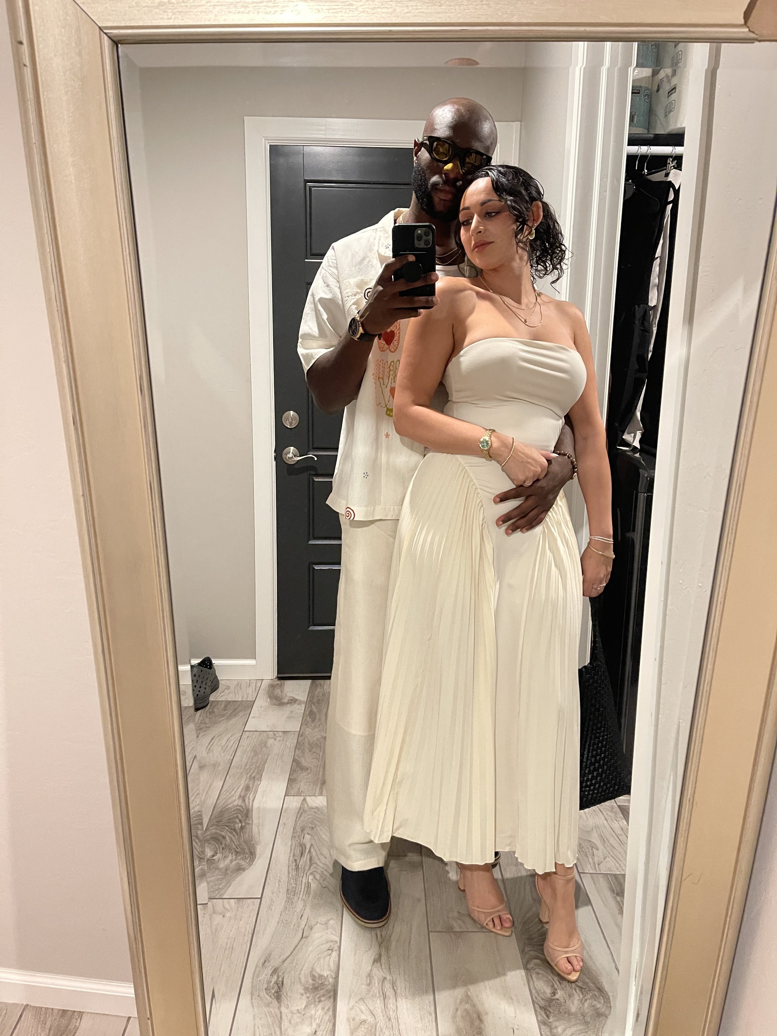 Couple taking a mirror selfie, dressed in coordinated white outfits, with the man wearing sunglasses and holding a smartphone.