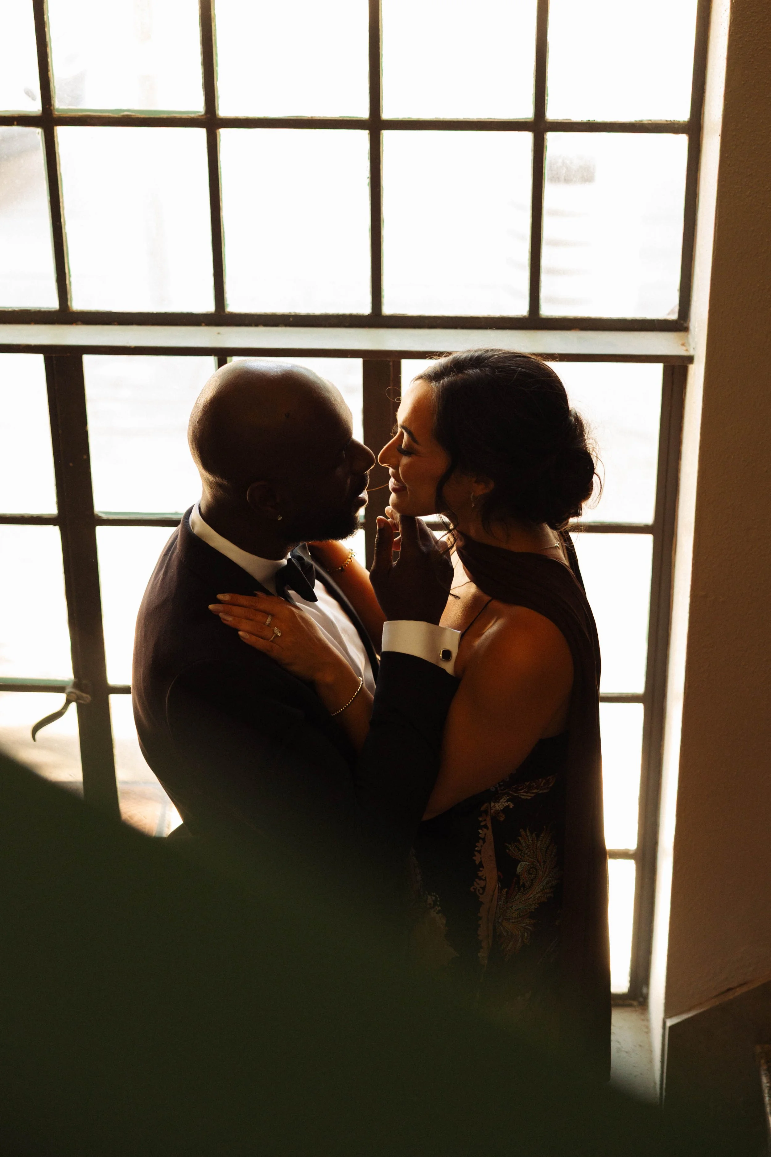 Bride and groom embracing and smiling nose-to-nose in front of a sunlit window
