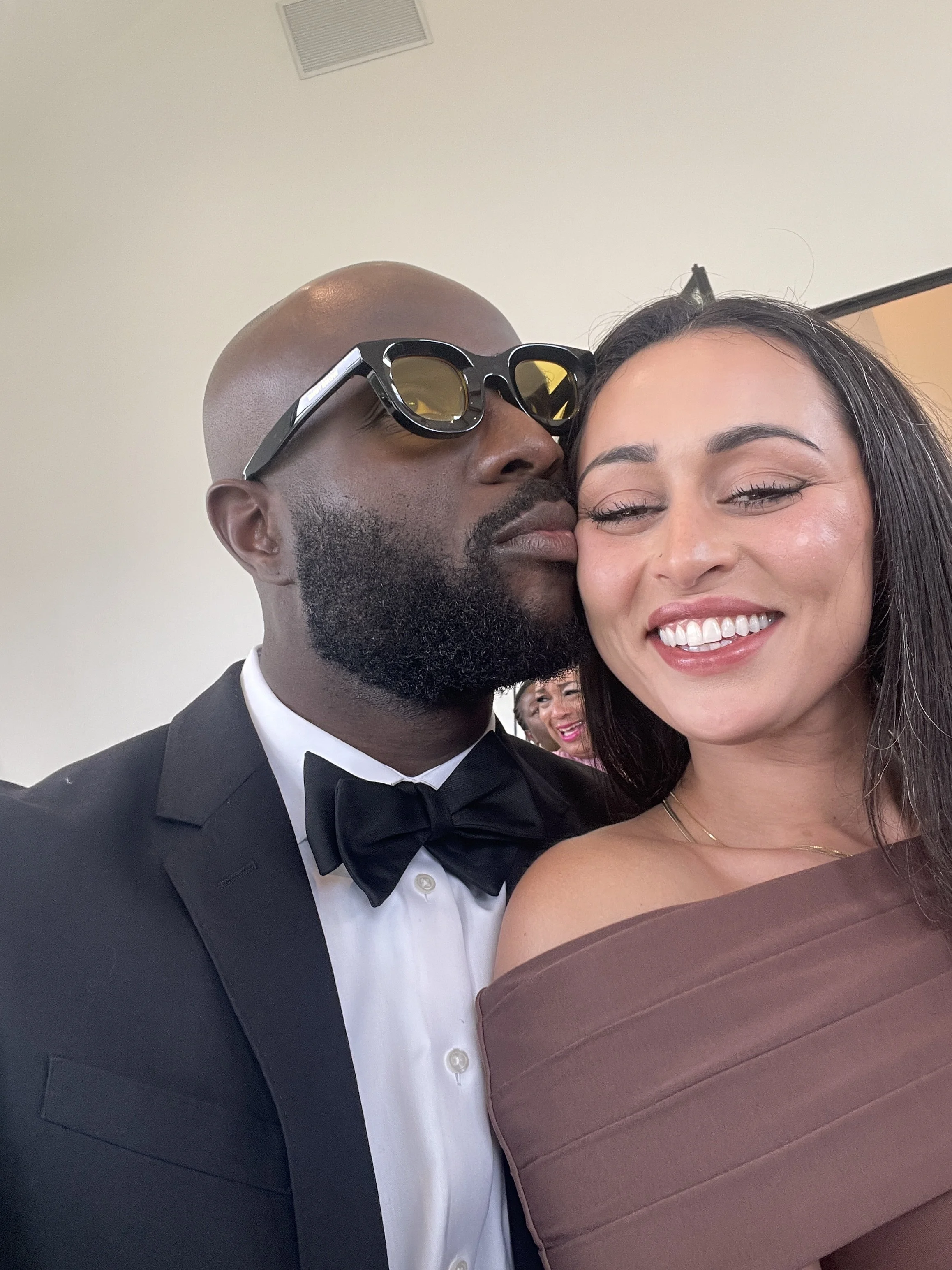 Smiling couple taking a close-up selfie at a formal event, with the man wearing sunglasses and a tuxedo and the woman in a mauve dress.
