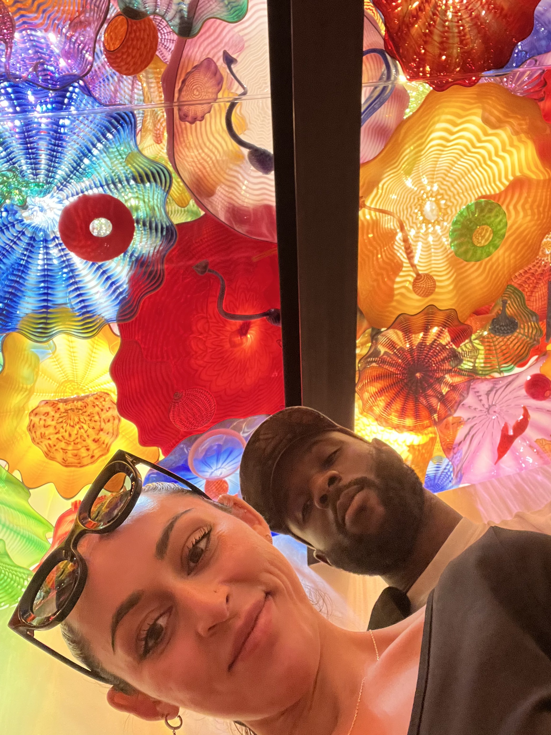 Two people pose in front of a colorful illuminated glass art installation filled with abstract flowers and swirling patterns.