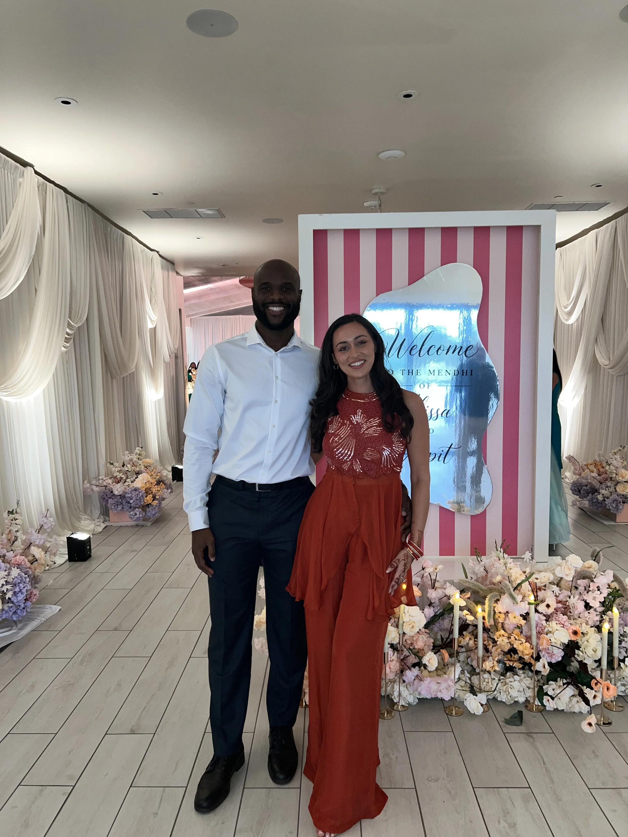 Couple posing at a colorful Mendhi celebration decorated with pink stripes, flowers, candles, and draped curtains.
