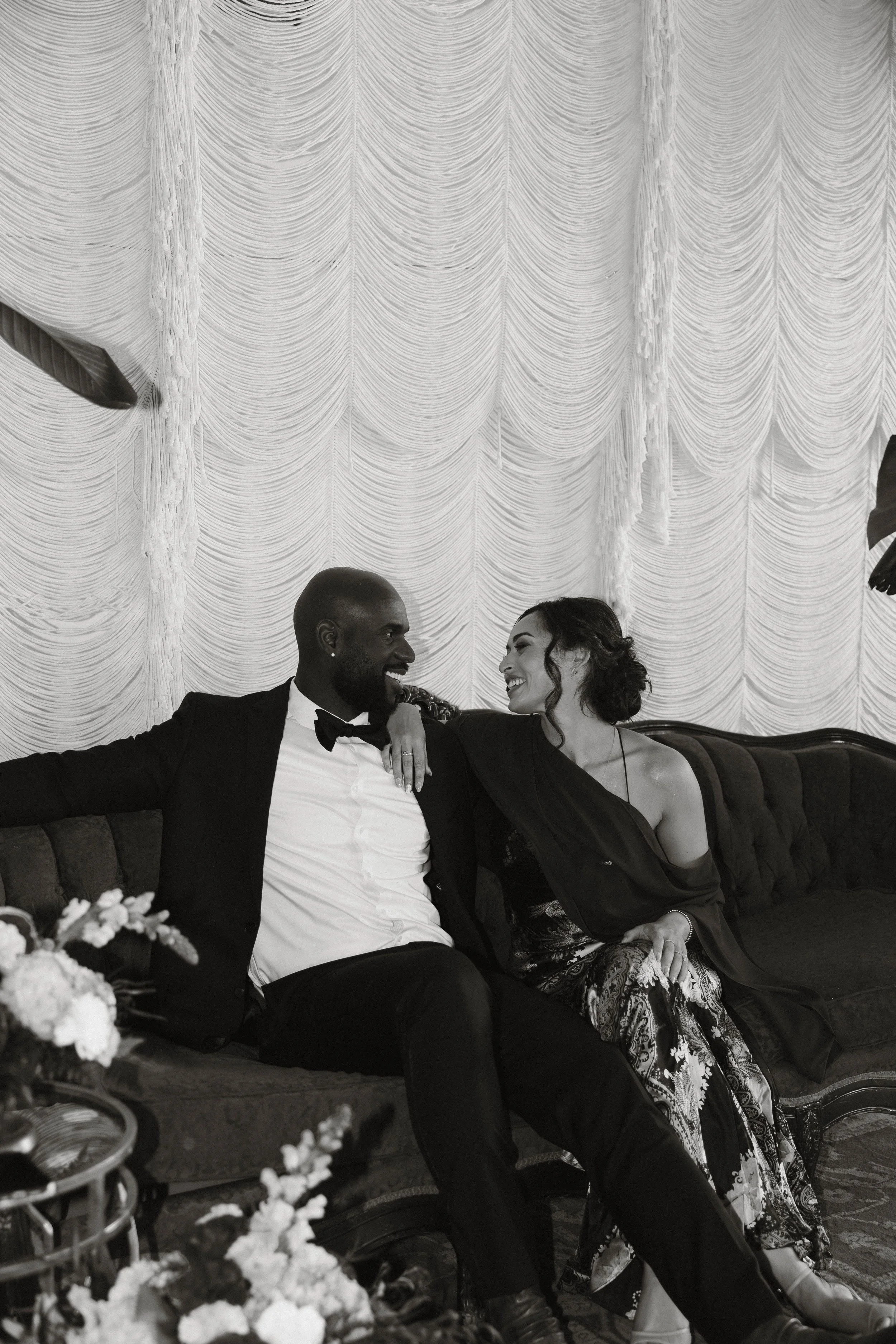 Black-and-white portrait of a smiling couple seated together on an ornate sofa in formal attire.