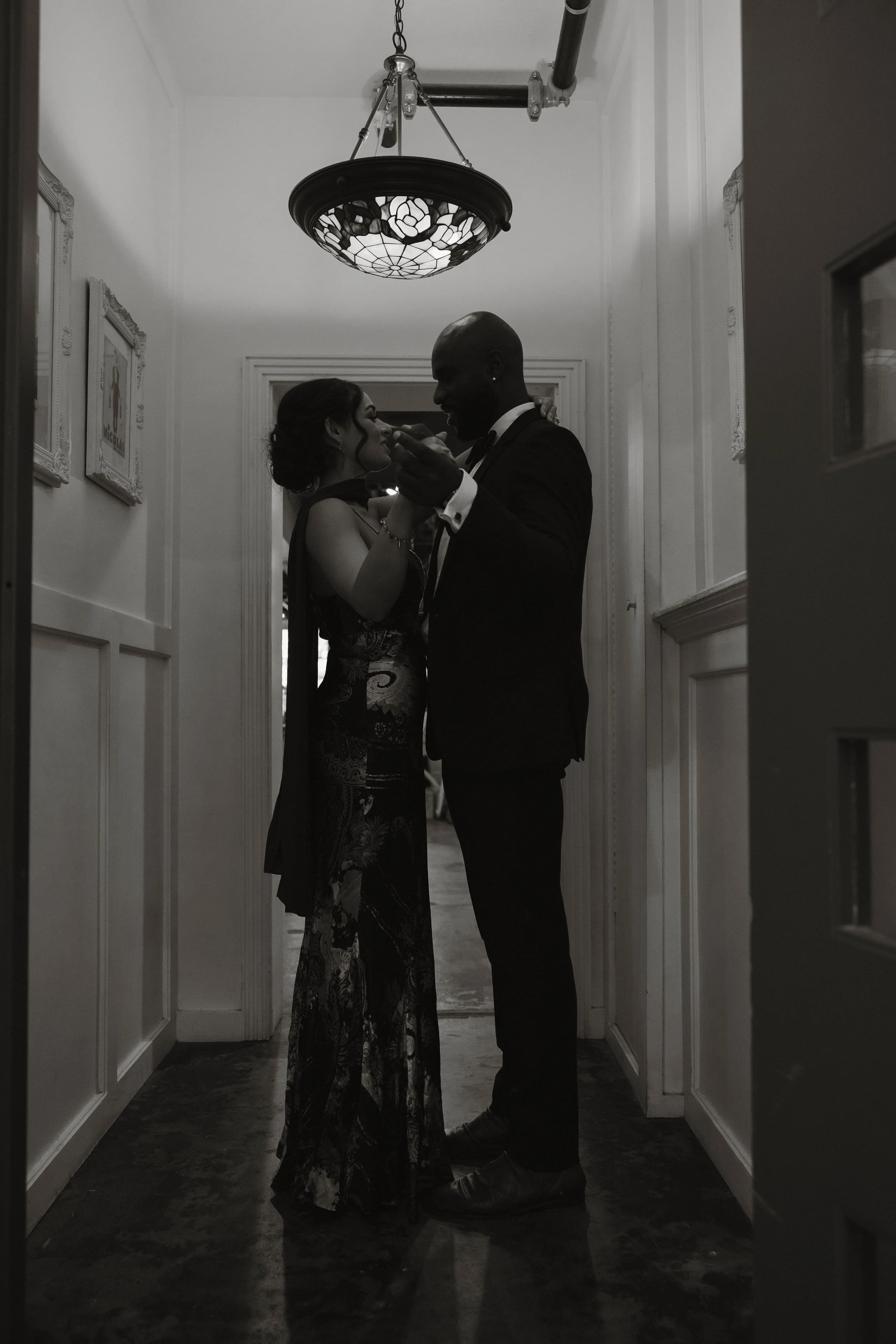 Black-and-white photo of a couple embracing beneath a stained-glass pendant light in an elegant hallway