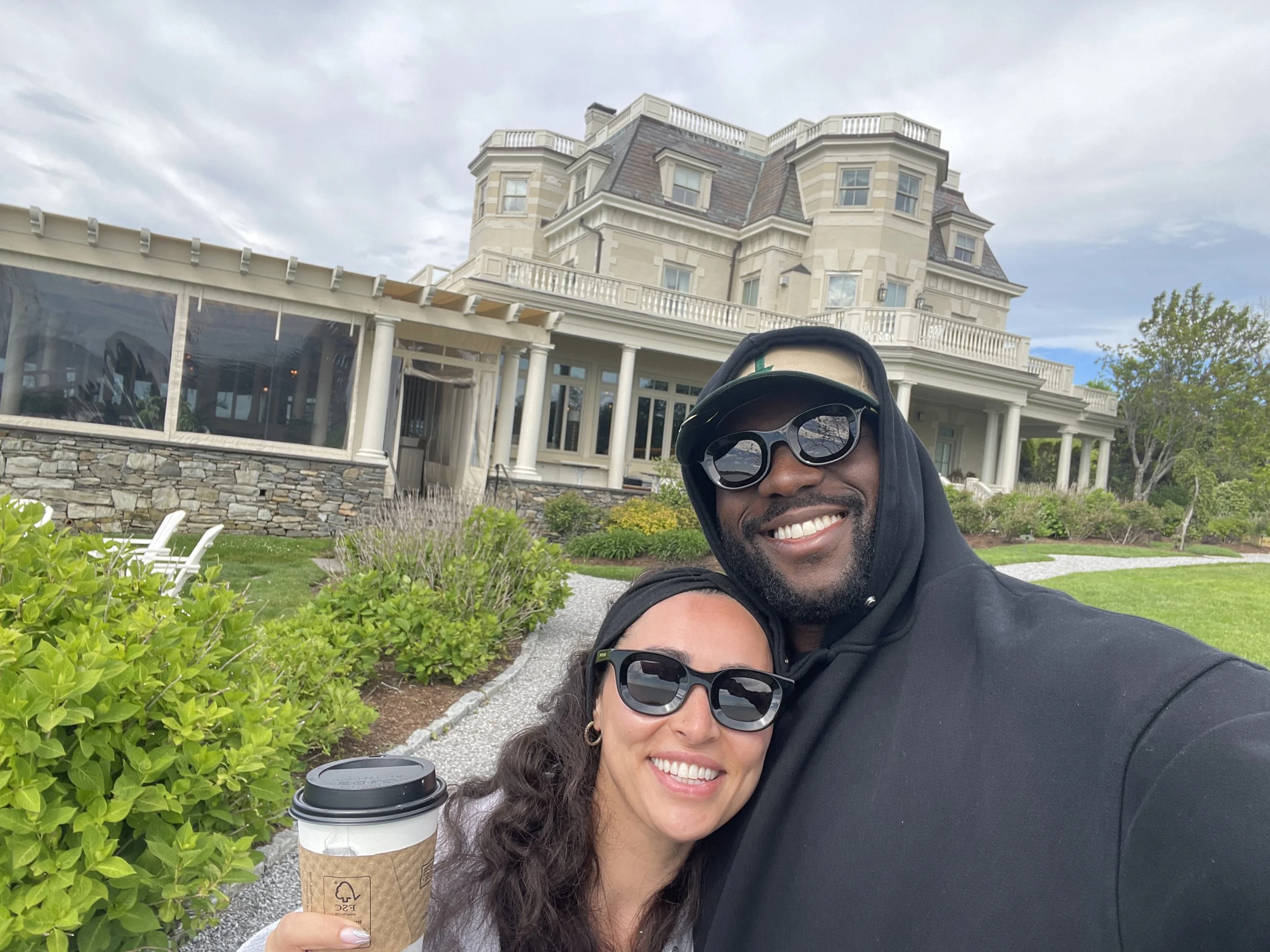 Smiling couple wearing sunglasses take a selfie outside a grand mansion, with one person holding a coffee cup.